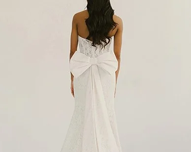 Back view of a woman in a white lace wedding dress with a large bow at the waist, standing against a plain white background.