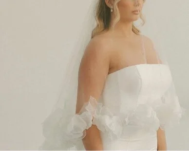 A woman in an elegant white dress with delicate spaghetti straps and ruffled sleeves, standing against a plain background. She has blonde hair styled in soft waves and is wearing earrings and a veil.