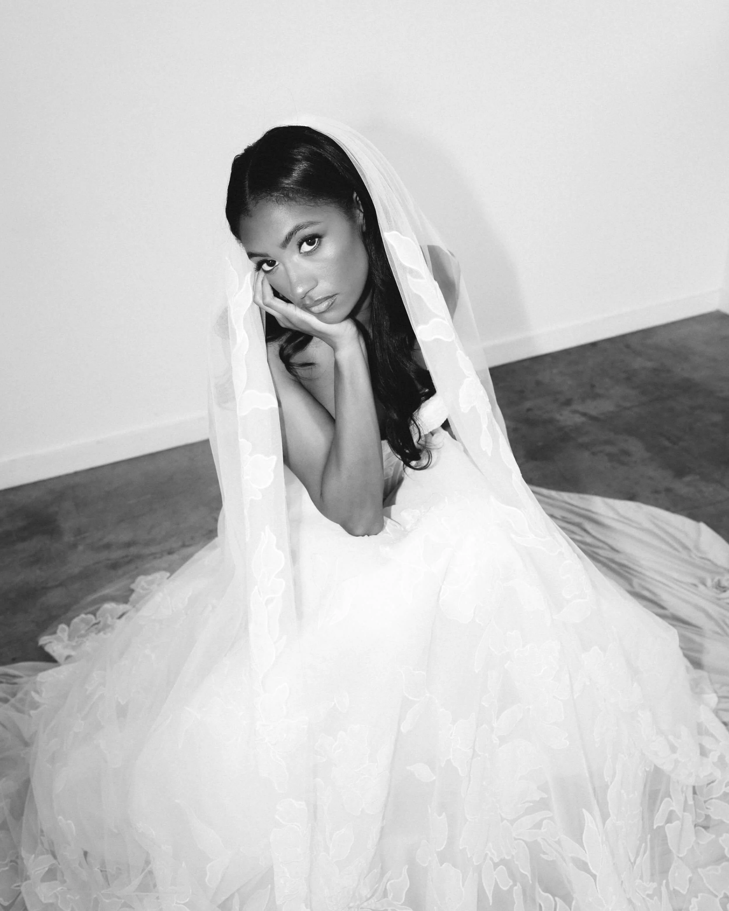 A woman in a wedding dress with a veil sitting on the floor, looking up at the camera with her hand resting on her face.