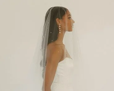 Side view of a woman in a wedding dress with a veil and pearl earrings, standing against a plain light background.