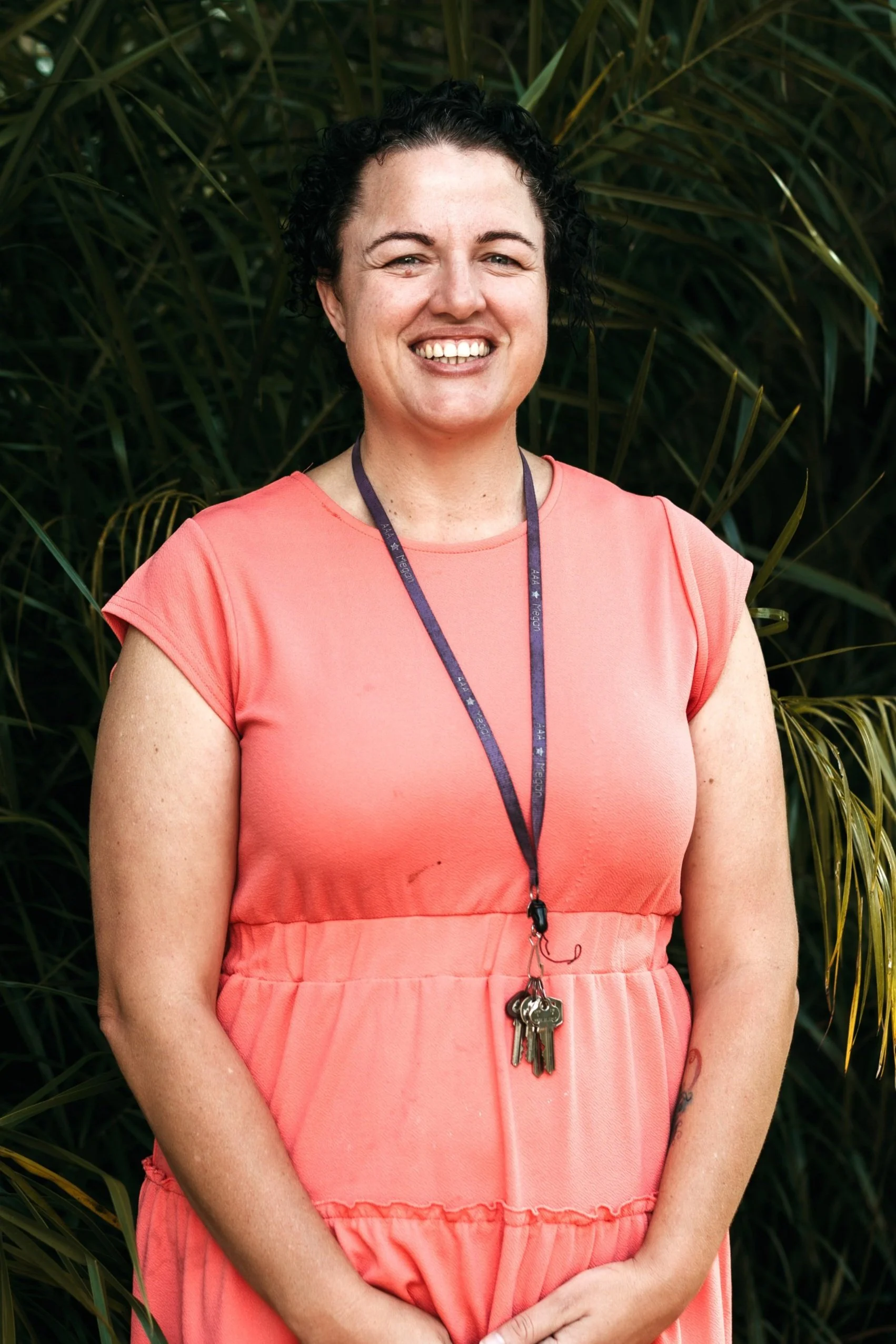 A woman with short, curly dark hair with blue highlights, smiling, wearing a teal dress and a blue lanyard with white text that reads "AAA Megan," standing outdoors in front of green foliage.
