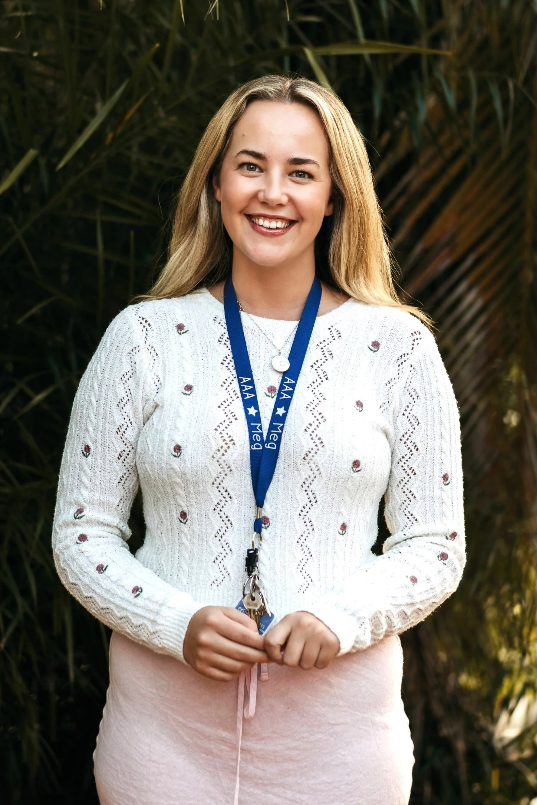 A young woman with blonde hair, wearing a light blue sleeveless top, standing outdoors with green plants in the background. She has a purple lanyard labeled 'AAA Mega' around her neck and is smiling.