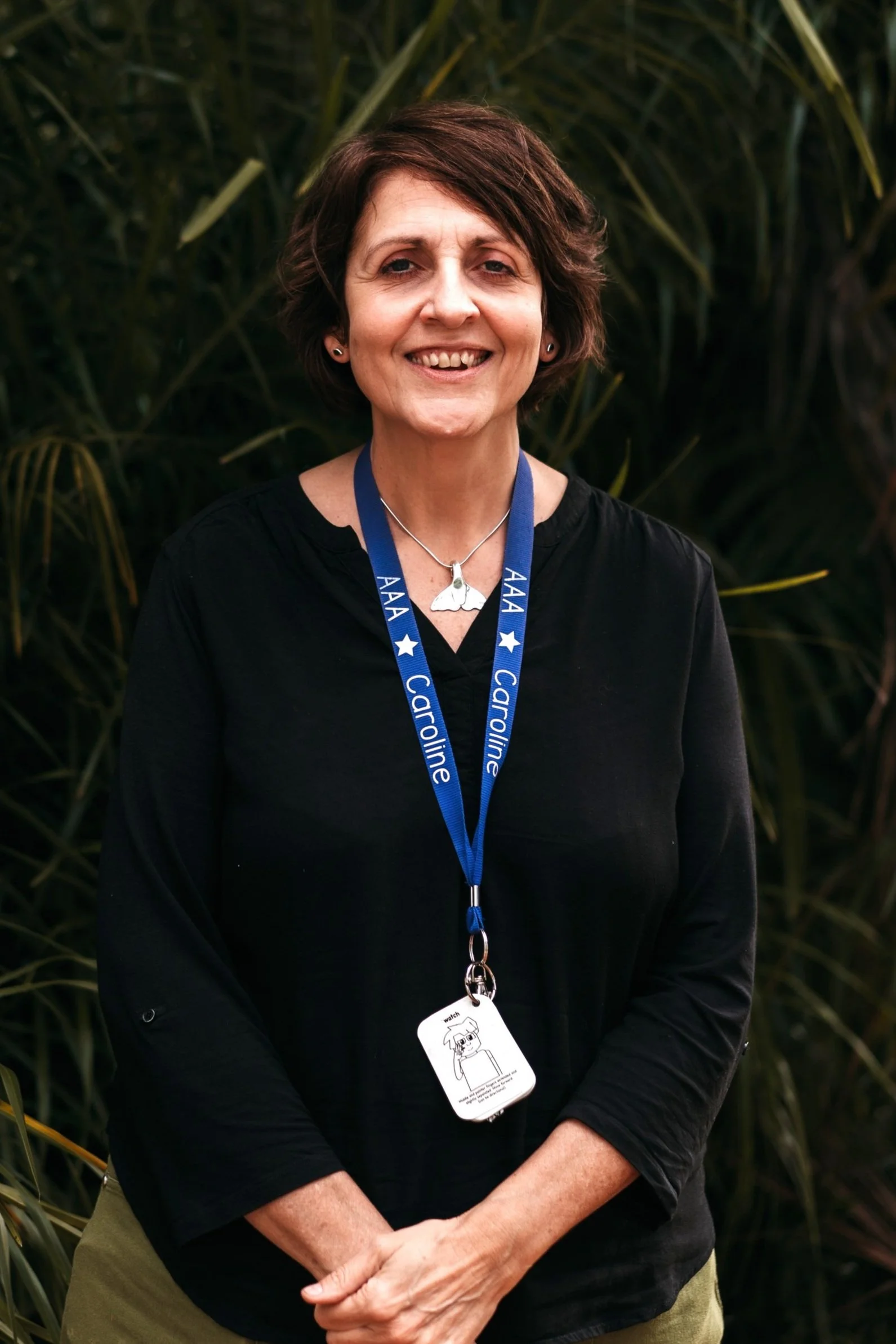 Smiling woman with short curly brown hair, wearing a black sleeveless top and a blue lanyard with white text and stars, stands outdoors with green foliage in the background.