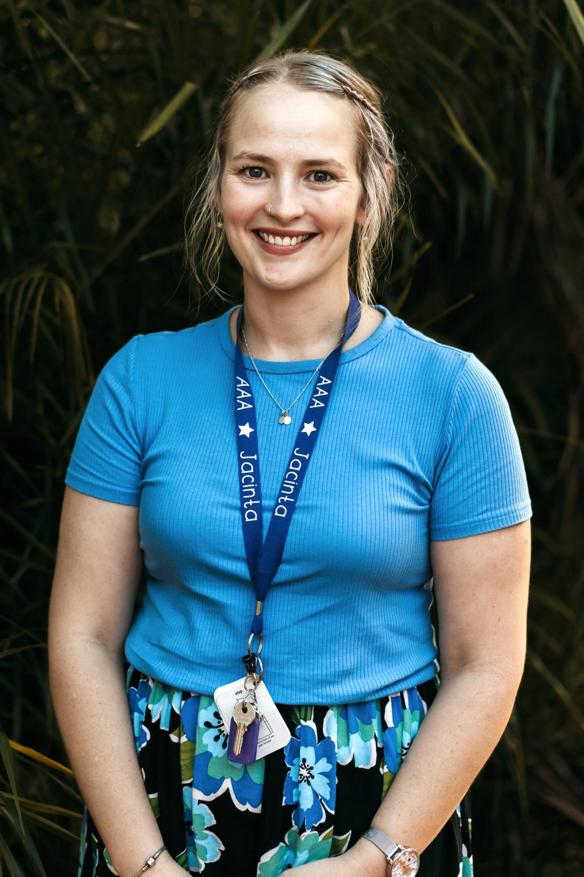 Young woman with blonde hair tied back, smiling, wearing a black and white checkered blouse and a blue graduation medal around her neck, standing outdoors with green foliage background.