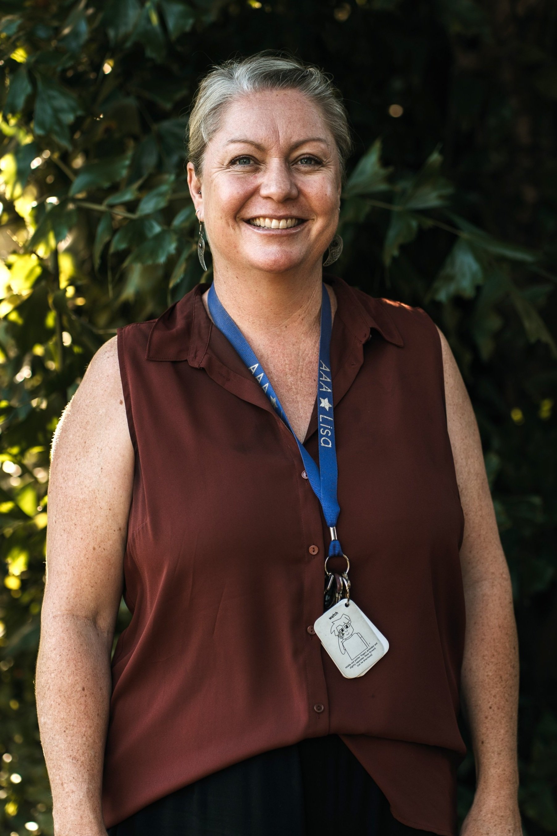 A woman with shoulder-length blonde hair smiling outdoors, wearing a blue blouse and a purple lanyard with the text 'AAA LISA' around her neck, standing in front of green foliage.
