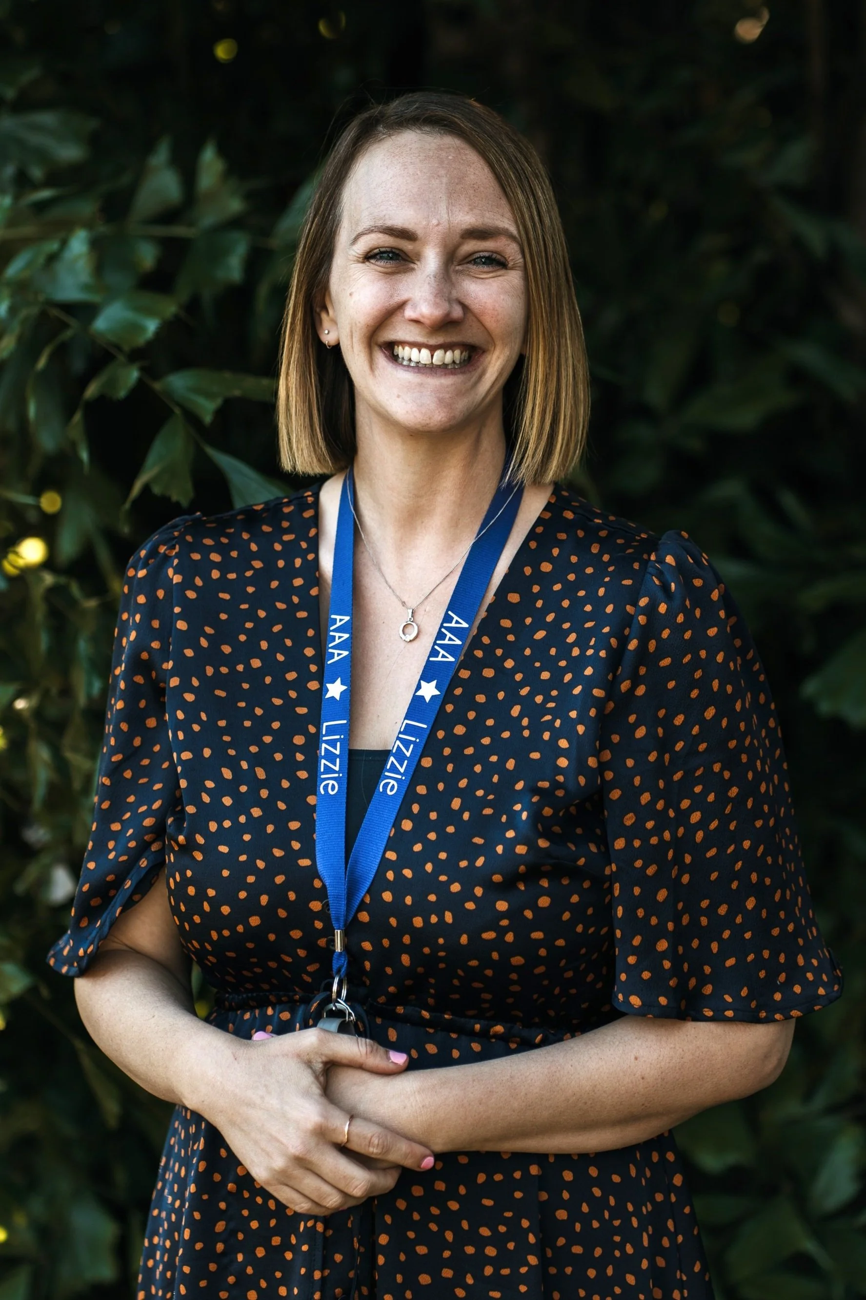 Smiling woman with blonde hair wearing a white top and a blue lanyard, standing outdoors with green foliage in the background.