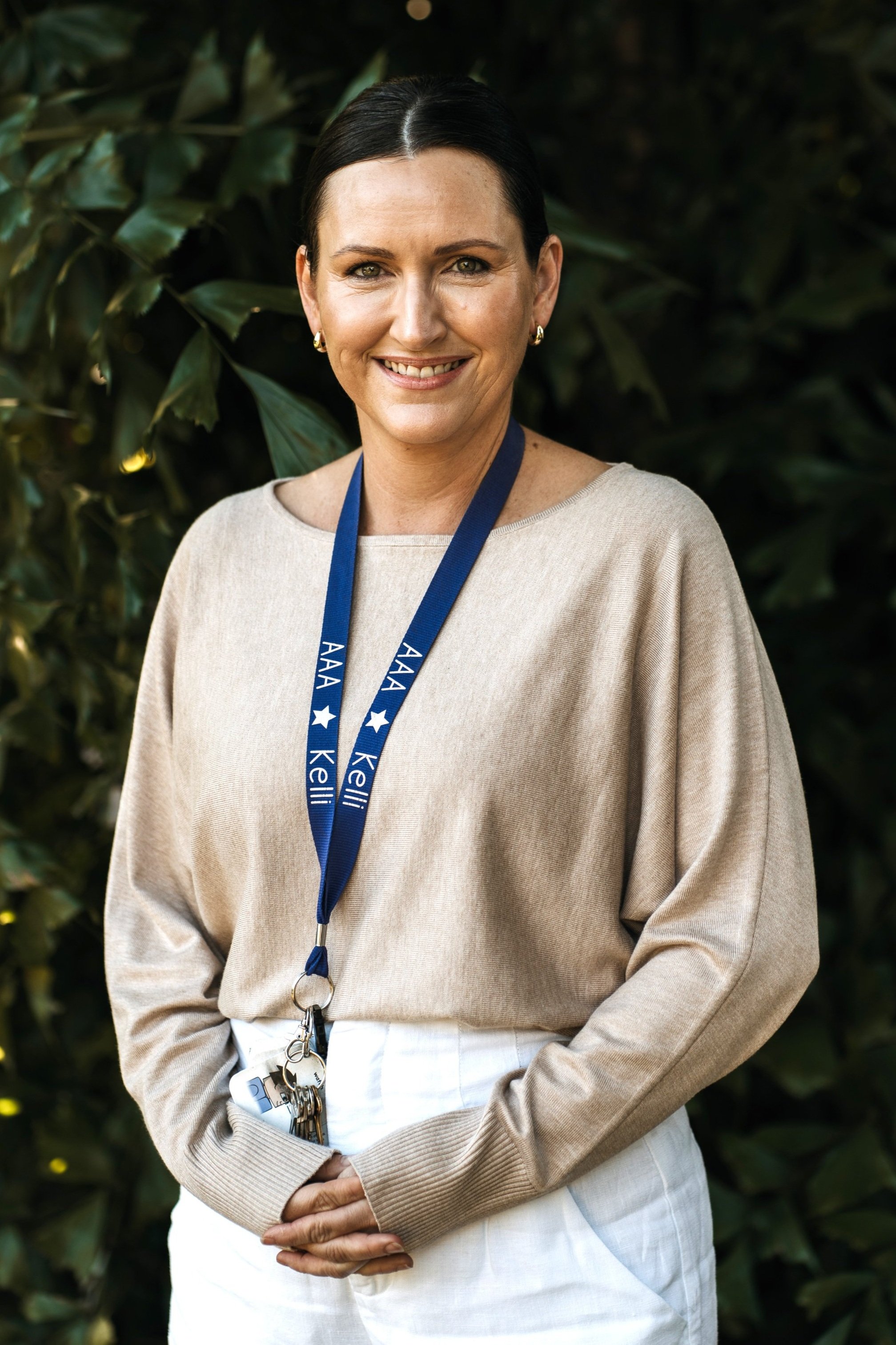 A smiling woman with shoulder-length brown hair, wearing a white shirt and a blue ALA medal lanyard, outdoors with green foliage in the background.