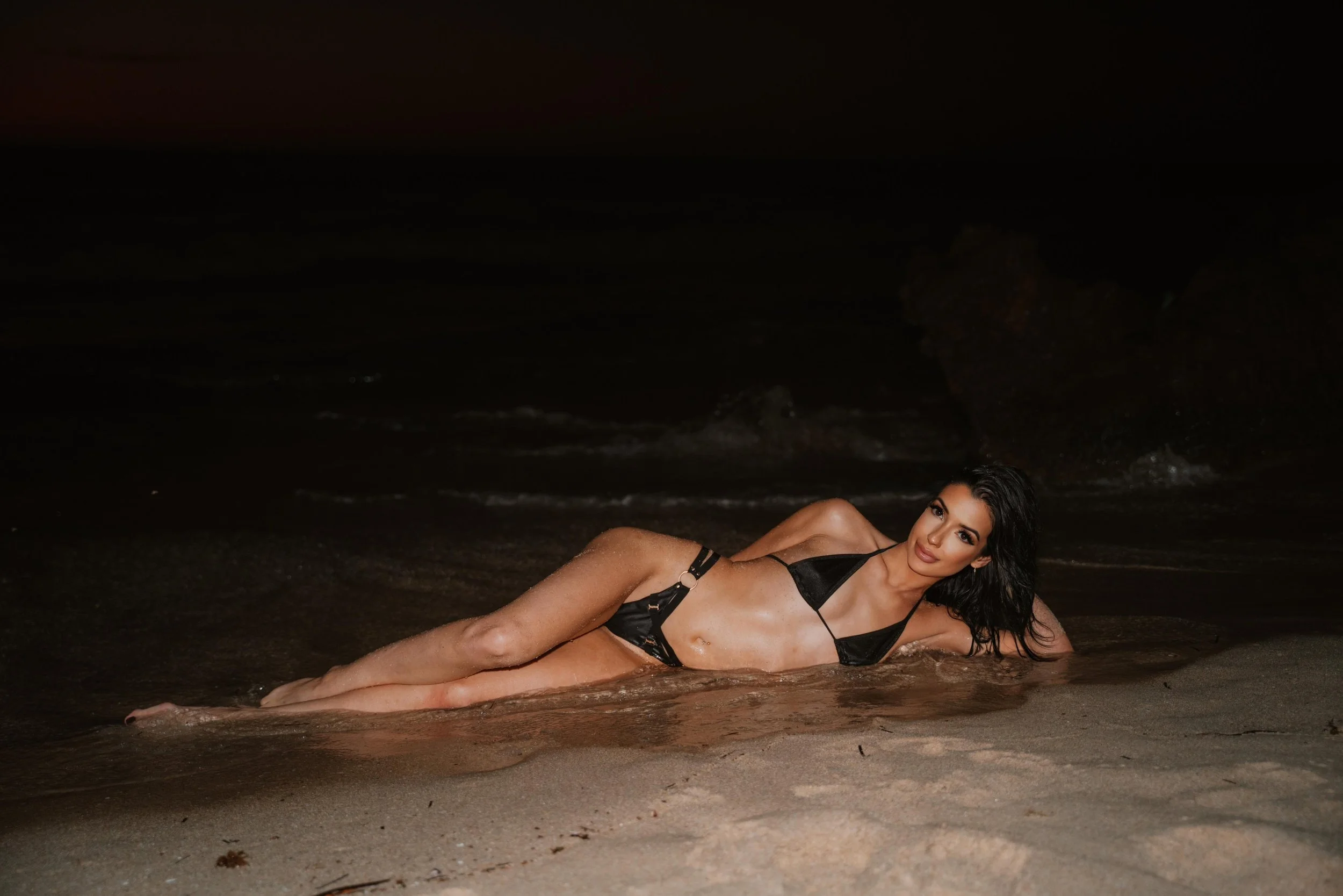 Woman lying on the beach at night, wearing black swimsuit, with wet hair and a confident expression.