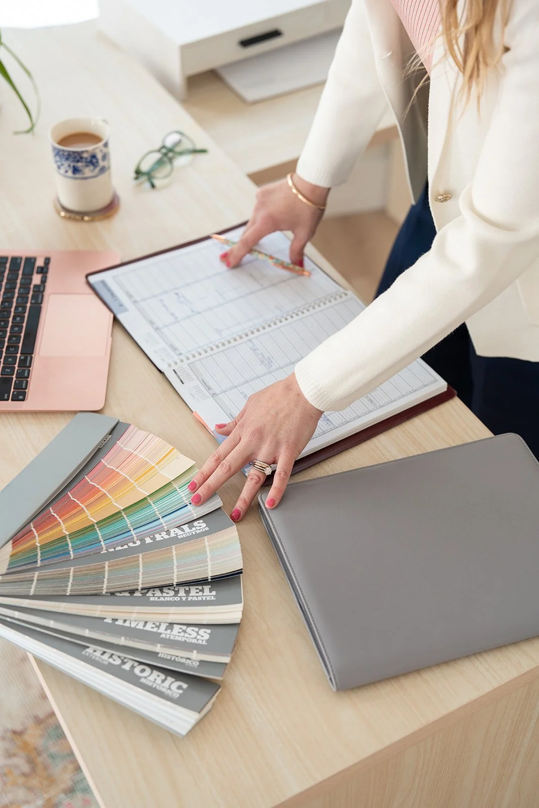Maura standing at her desk looking over a pantone color book, with appointment book, glasses & laptop on desk.