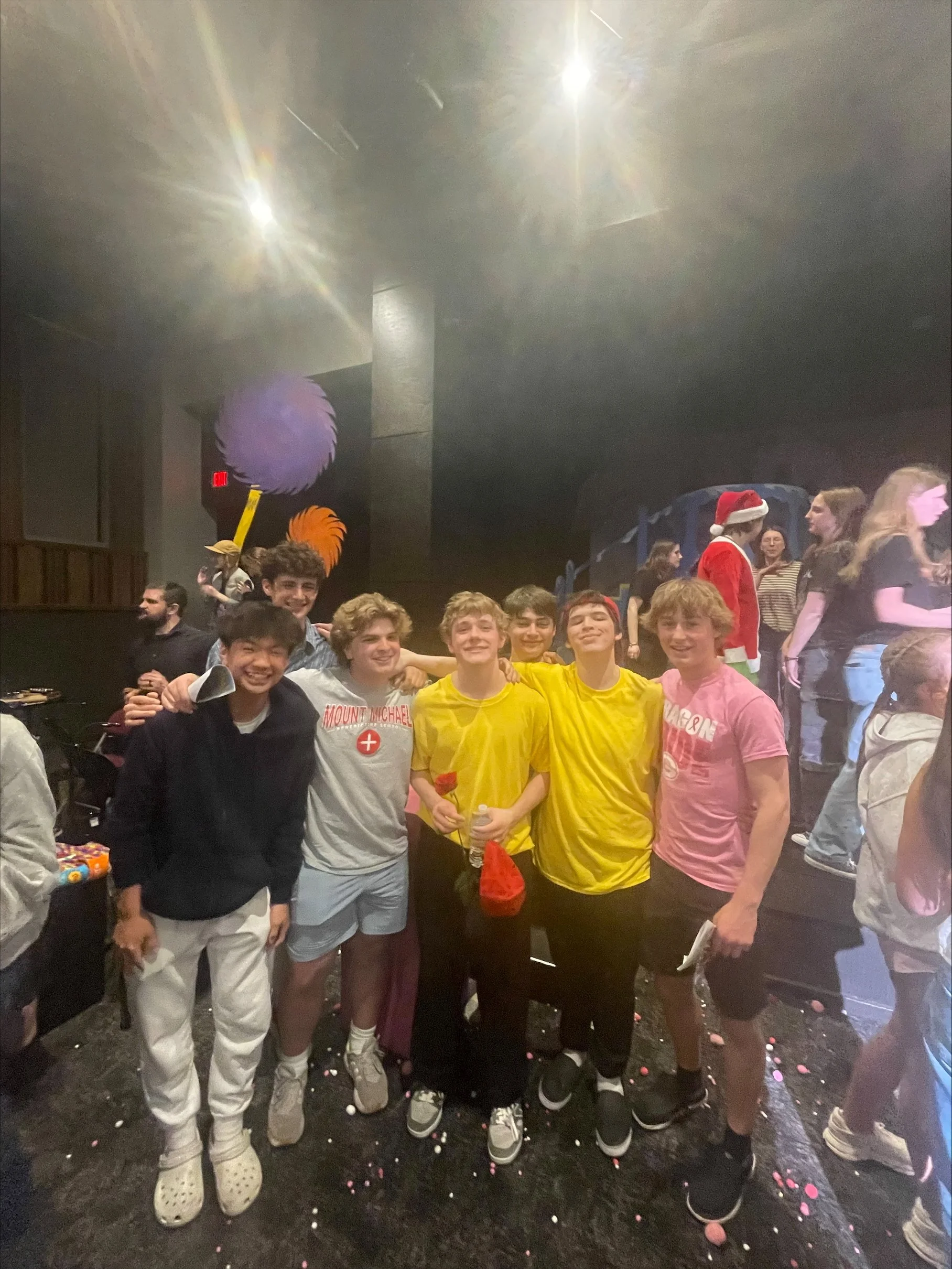 Group of young people posing indoors at an event, some wearing costumes and holding props, with a festive atmosphere.