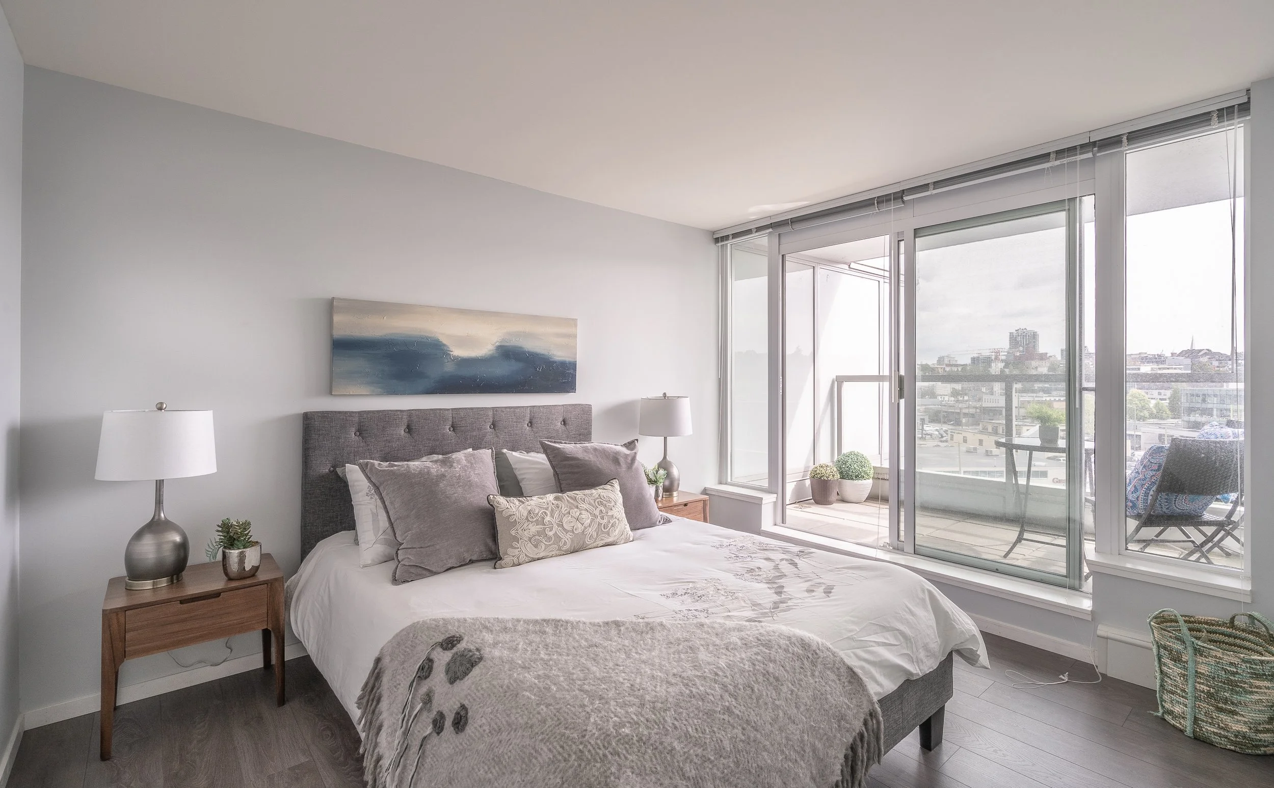 Bedroom with bed, two bedside tables with lamps, sliding glass door leading to balcony with outdoor furniture and city view