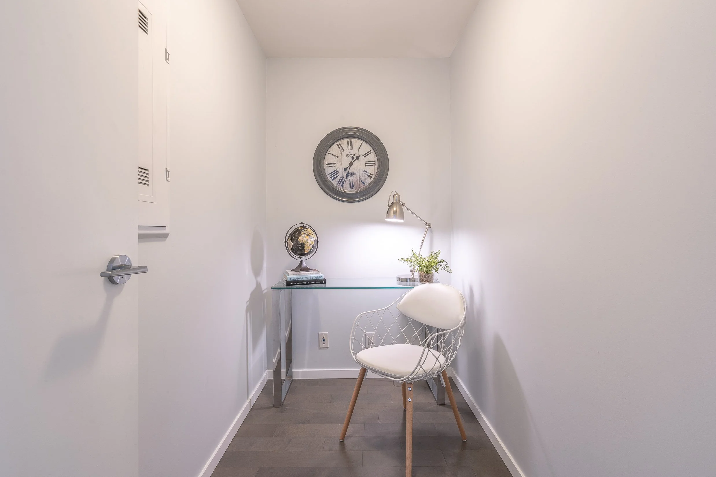 A minimalist home office with white walls, a glass desk, a white chair with wooden legs, a black and white globe, a stack of books, a plant, a modern silver desk lamp, and a wall clock with Roman numerals.