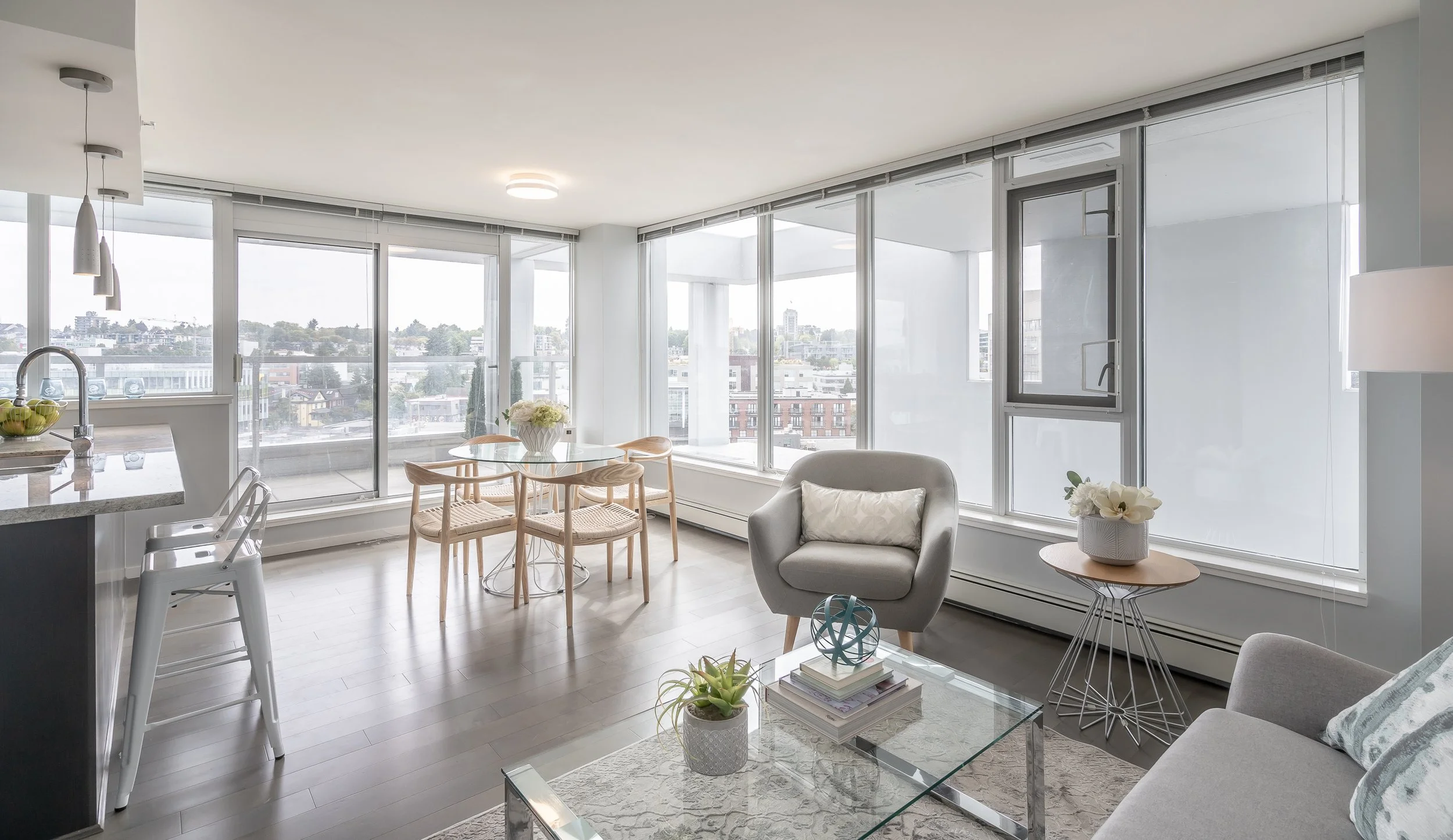 Modern living room with large windows, gray furniture, glass coffee table, round dining table with wooden chairs, and a kitchen counter with bar stools.