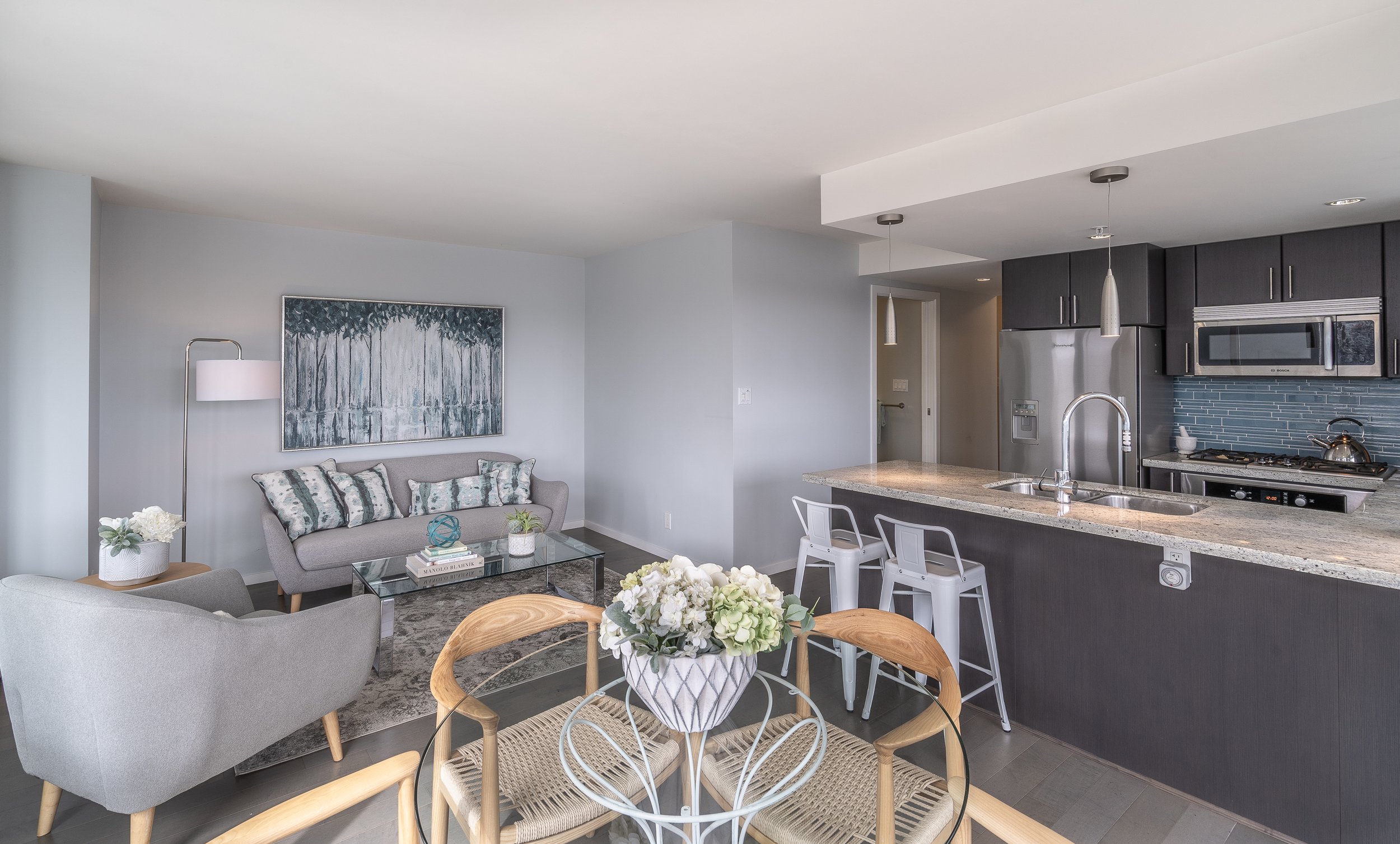 Modern open-plan kitchen and living room with gray sofa, glass coffee table, bar stools, dining table with flowers, stainless steel appliances, and blue tile backsplash.