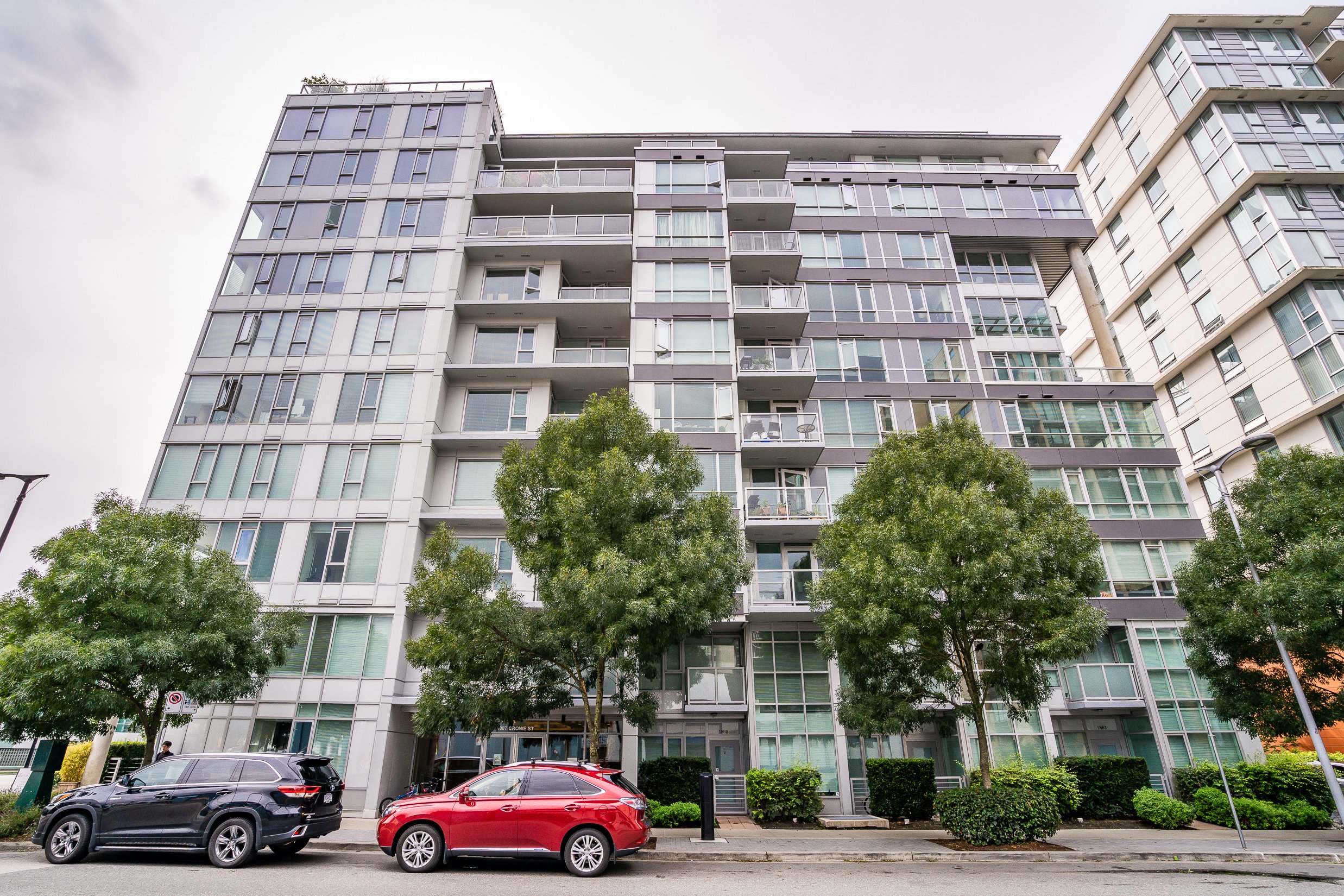 Front view of a modern multi-story apartment building with glass windows and balconies, trees lining the sidewalk, and parked cars in front.