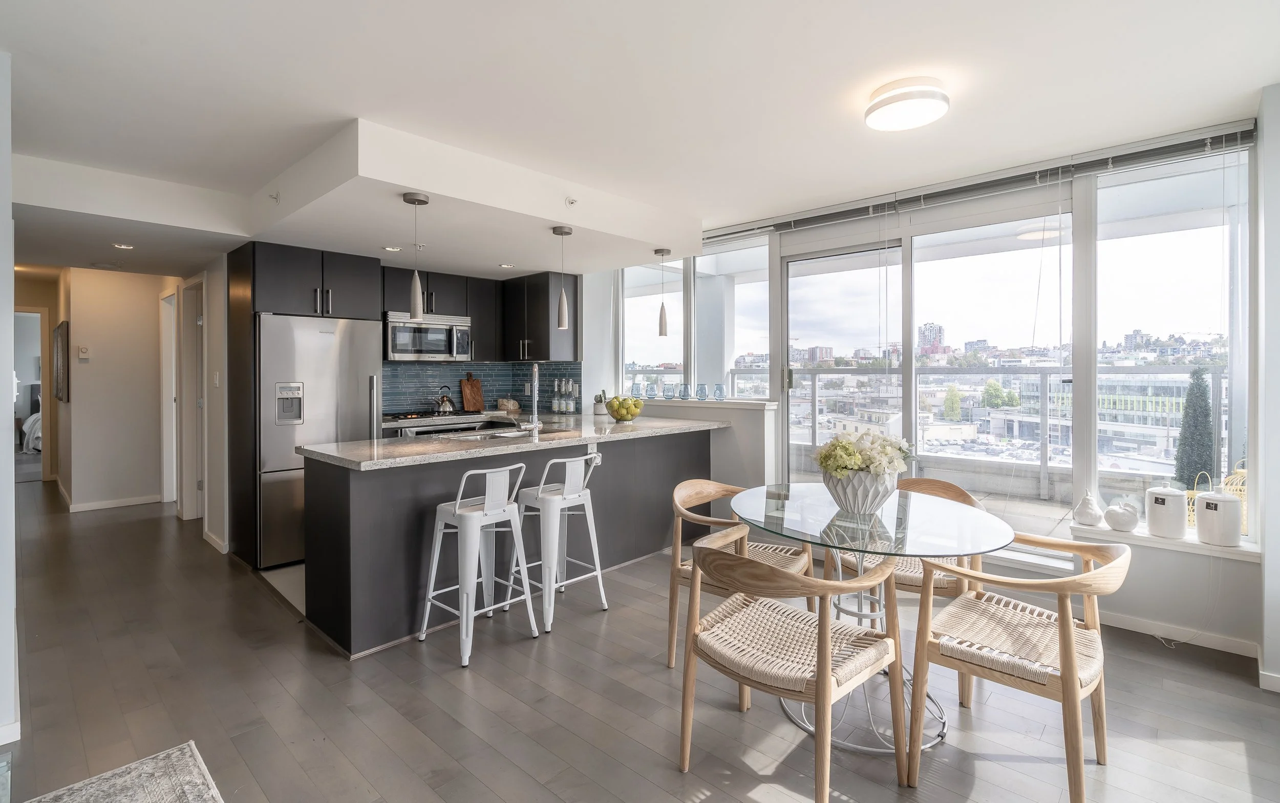 Modern kitchen and dining area with dark cabinetry, stainless steel appliances, a glass dining table with wooden chairs, and large floor-to-ceiling windows overlooking a cityscape.