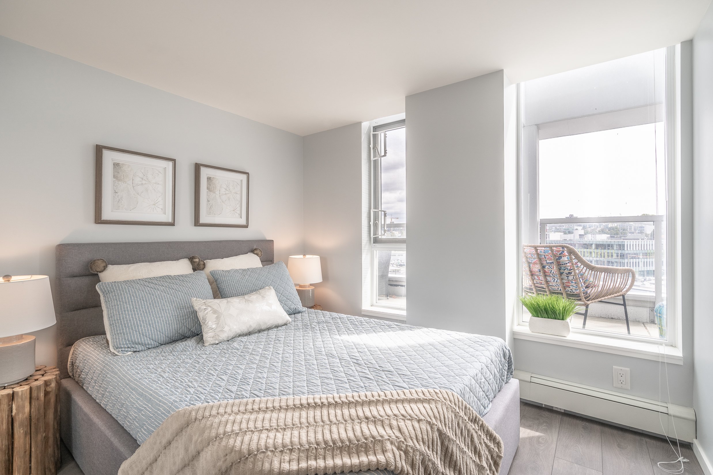 Modern bedroom with gray upholstered bed, decorative pillows, two nightstands with lamps, Canadian flag patterned outdoor chair, window with city view, potted grass, and framed artwork.