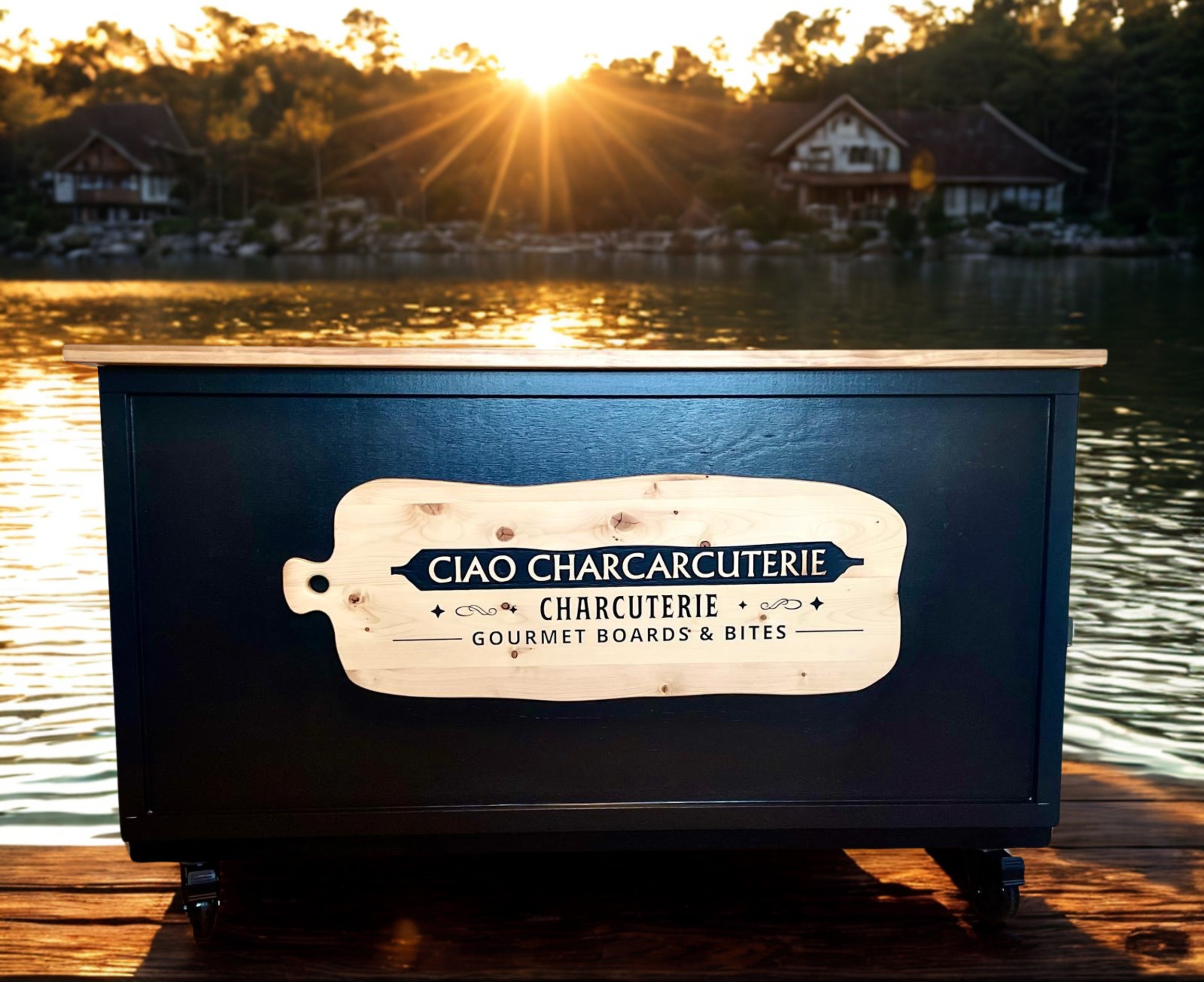 A black wooden cart with a light wood sign that reads 'CIAO CHARCUTERIE, CHARCUTERIE, GOURMET BOARDS & BITES,' positioned on a wooden dock by a body of water during sunset. In the background, there are two house-like structures and trees, with the sun setting behind the trees.