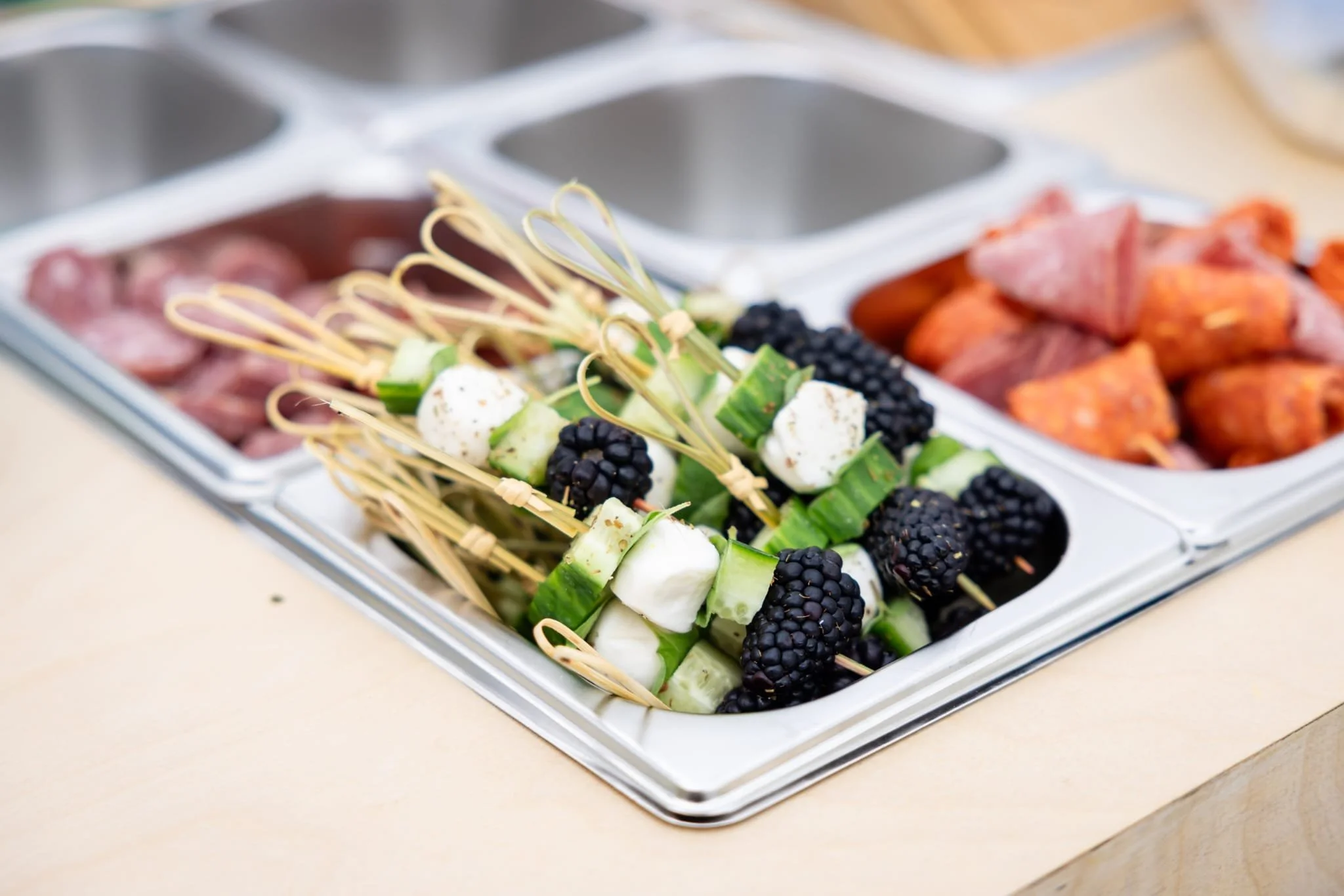Skewers with cucumber, mozzarella, and blackberries in a buffet tray, with other food items in the background.