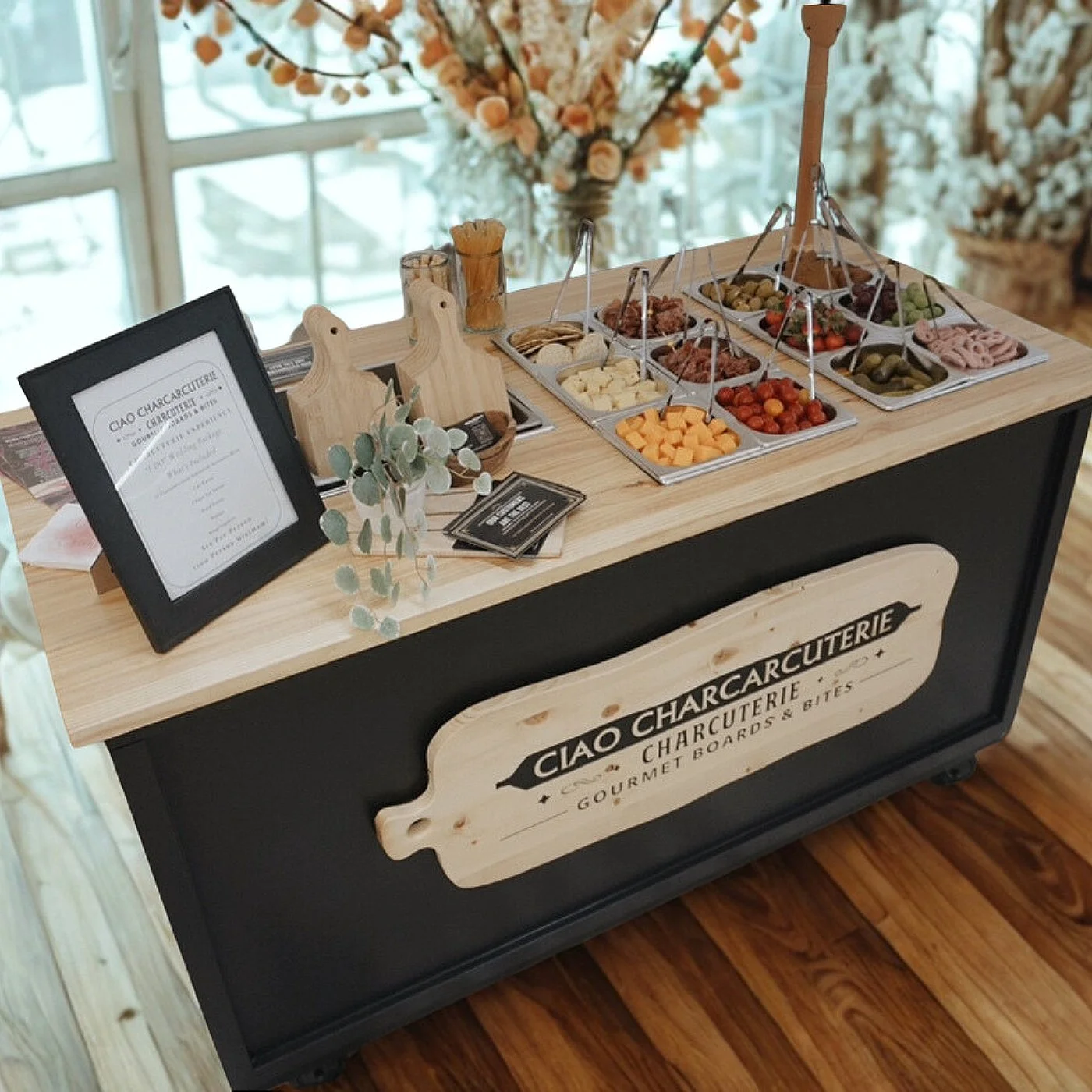 A charcuterie board stand with various cheeses, meats, olives, and vegetables on a wooden table. There is a sign for "Ciao Charcuterie" and a menu or informational card. Decorative greenery and a large flower arrangement are in the background.