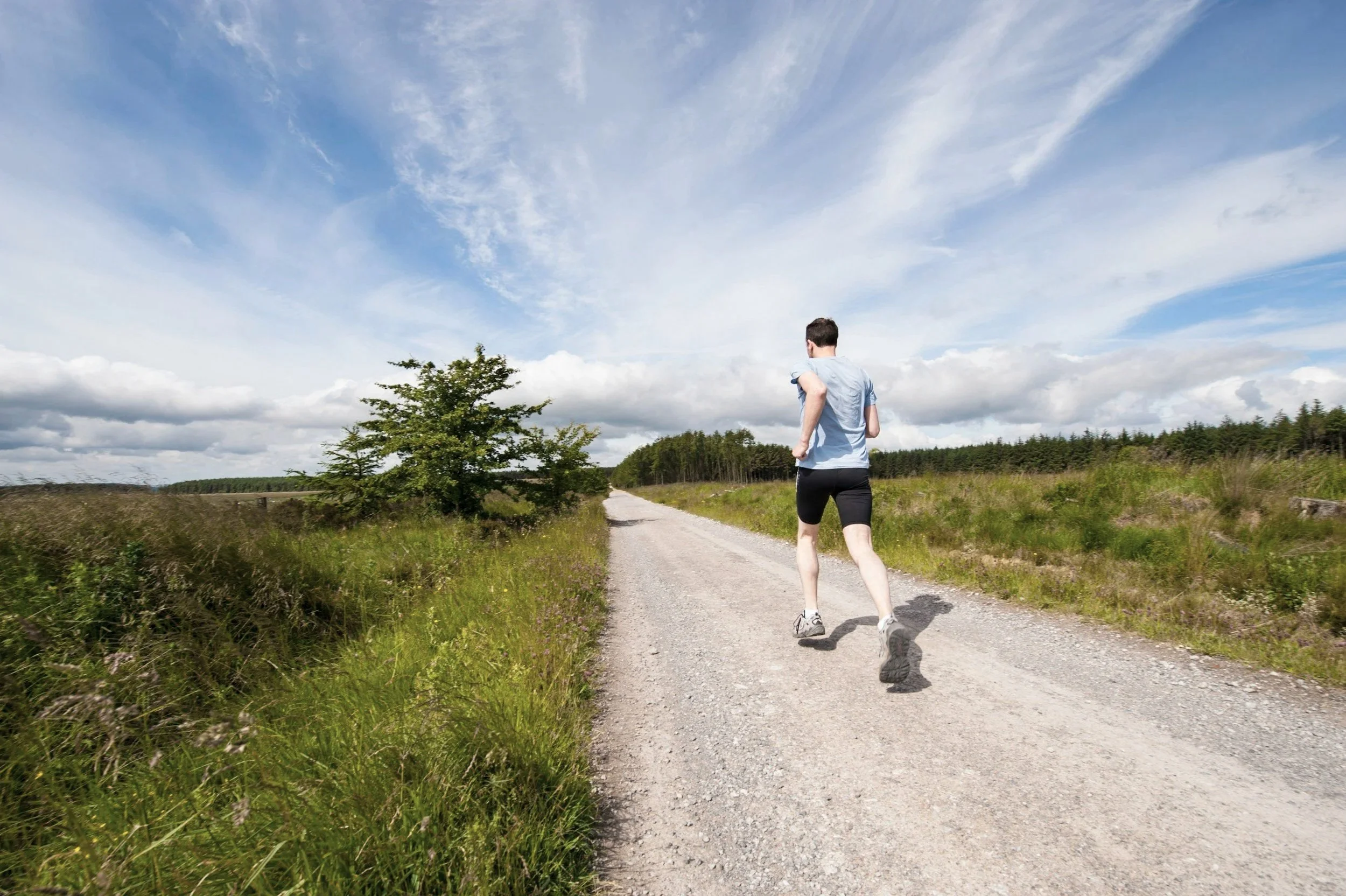 A person jogging on a gravel path in a grassy rural area under a partly cloudy sky.