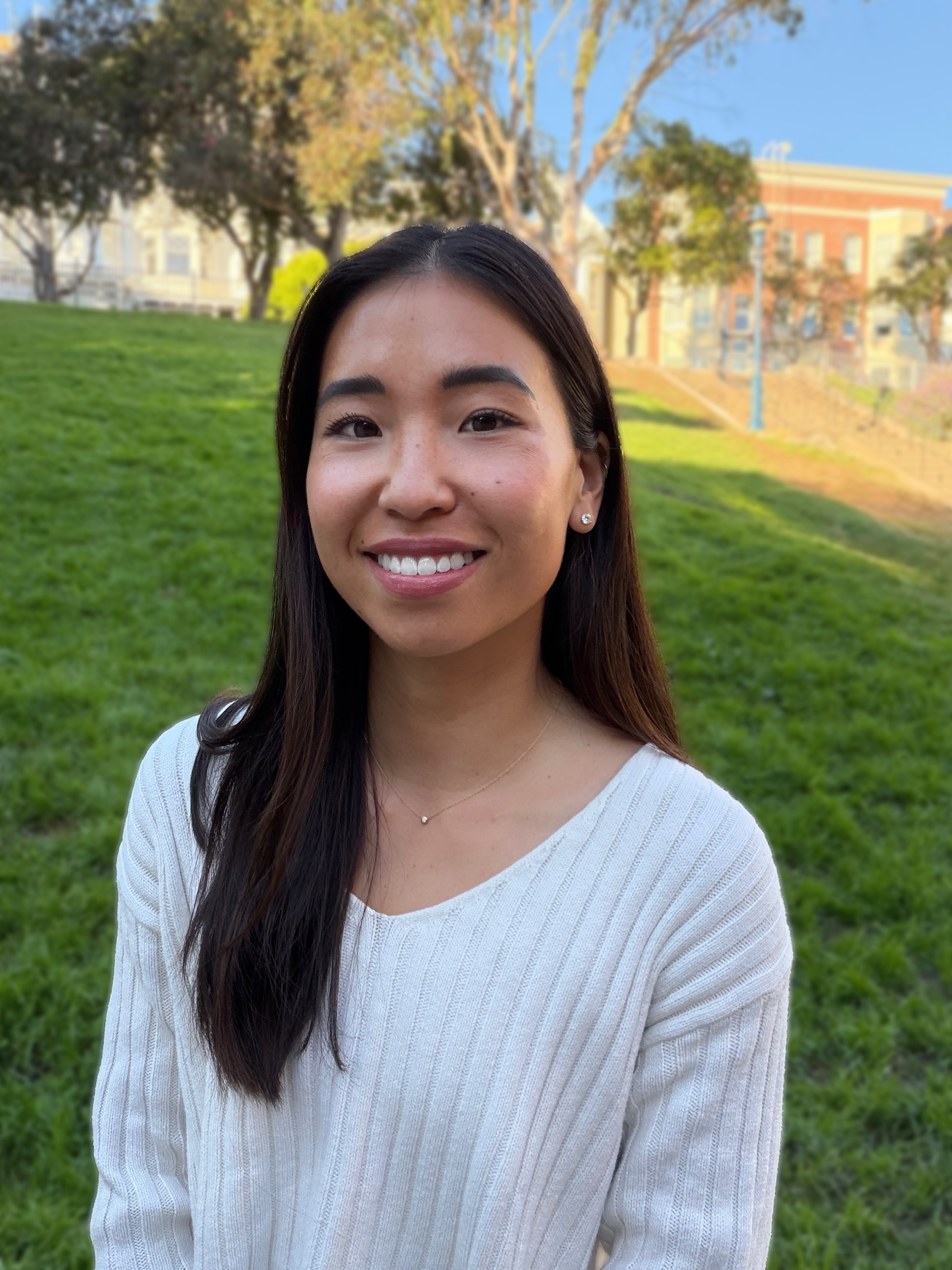 A young woman with long dark hair smiling outdoors on a grassy area with trees and buildings in the background.