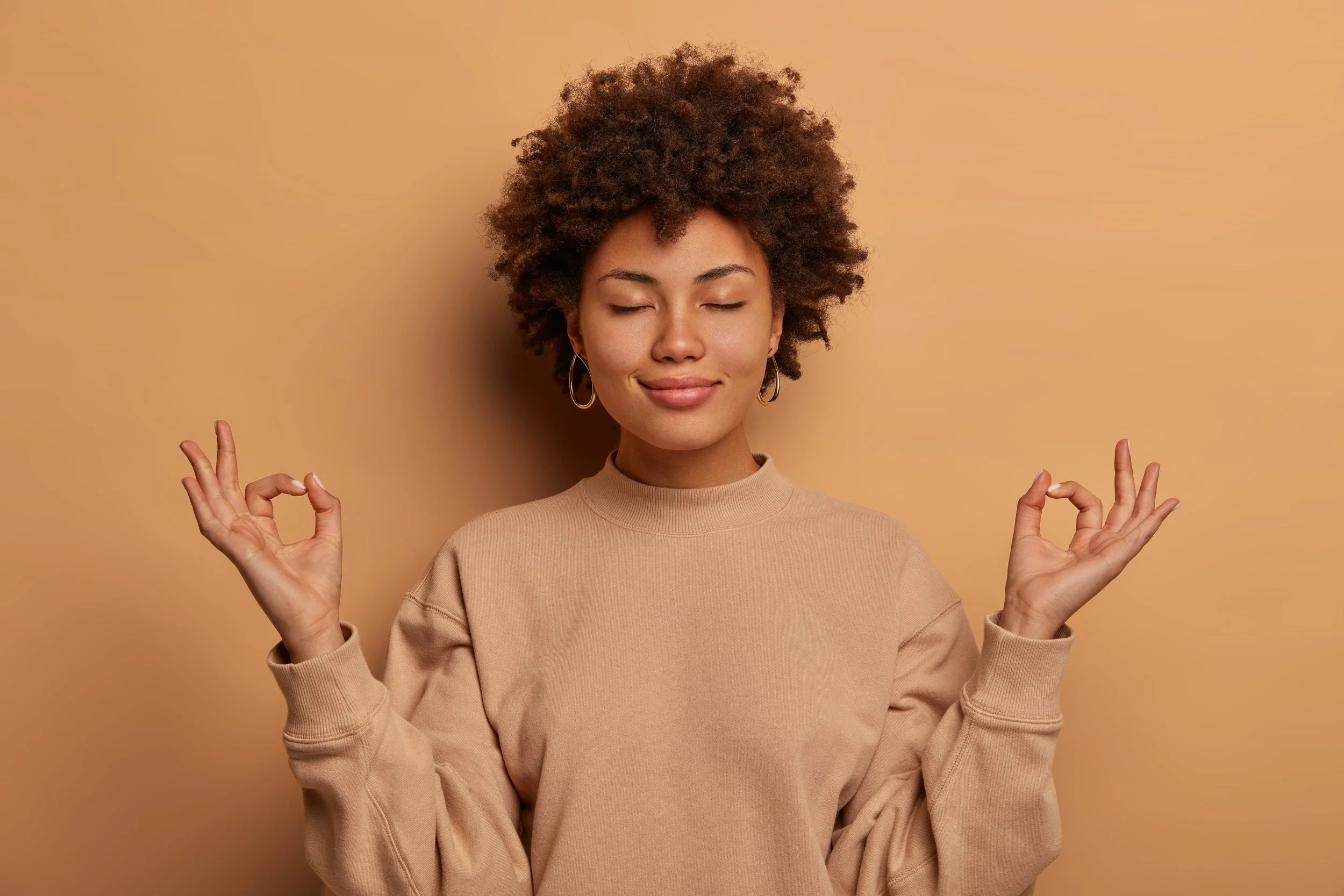 calm-relieved-dark-skinned-woman-takes-deep-breath-keeps-hands-sideways-zen-gessture-reaches-nirvana-practices-yoga-stands-with-closed-eyes-stands-stress-free-against-brown-wall.jpg