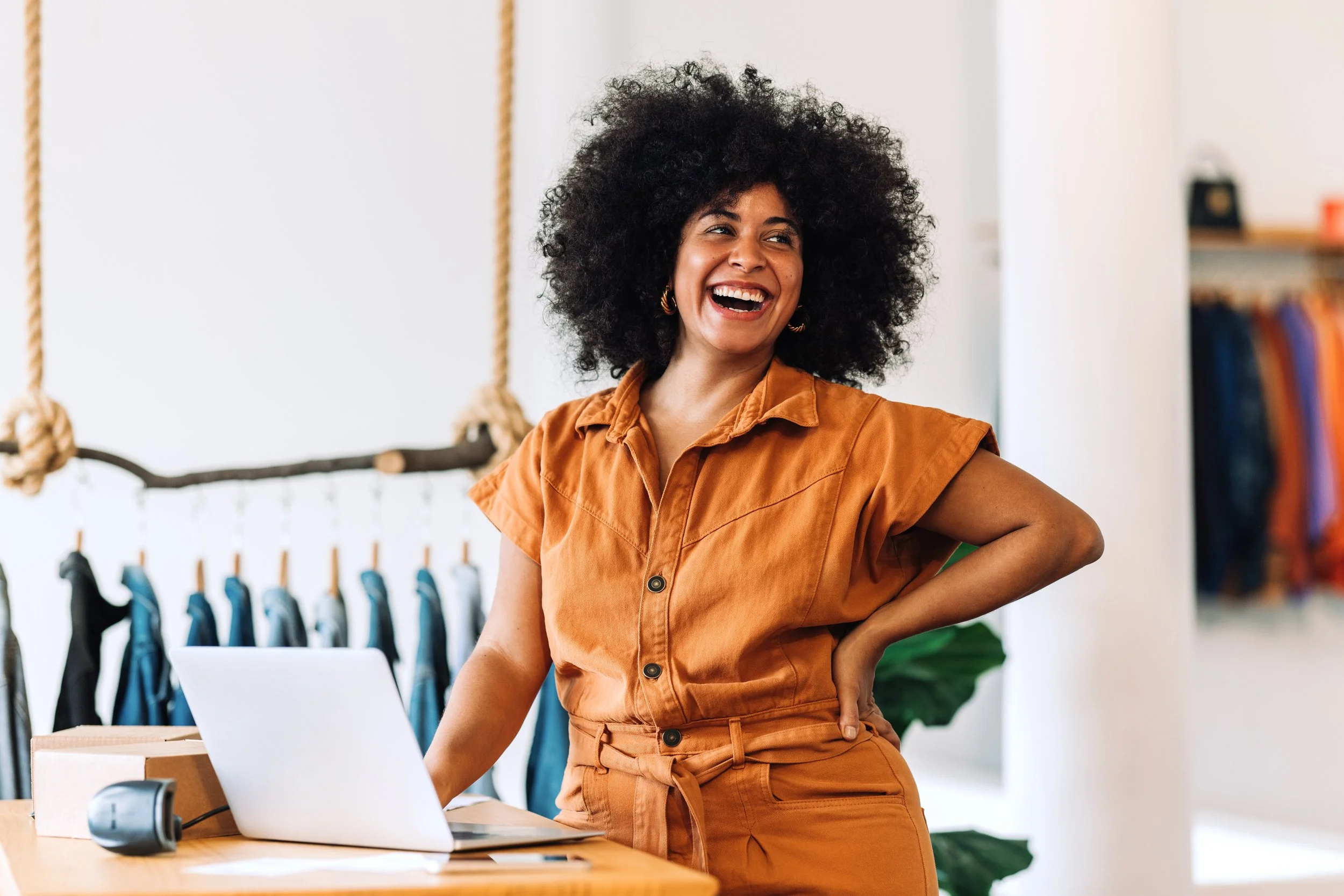 Woman with curly hair smiling and standing at a workspace with a laptop, headset, and cardboard box, in a modern room with a hanging clothing rack and books.