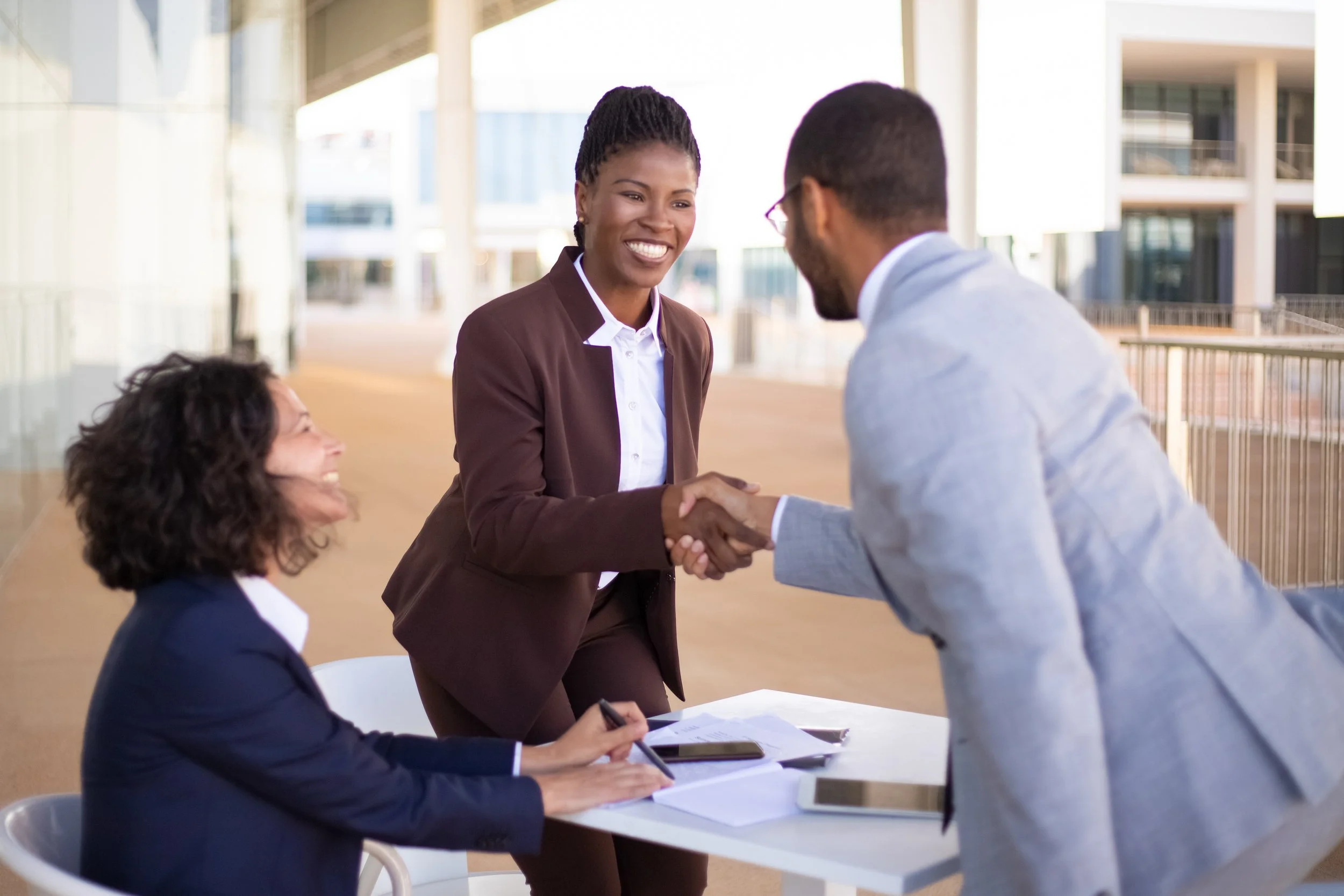 A woman in a brown suit shaking hands with a man in a gray suit, while another woman in a navy blazer smiles and takes notes at a table.