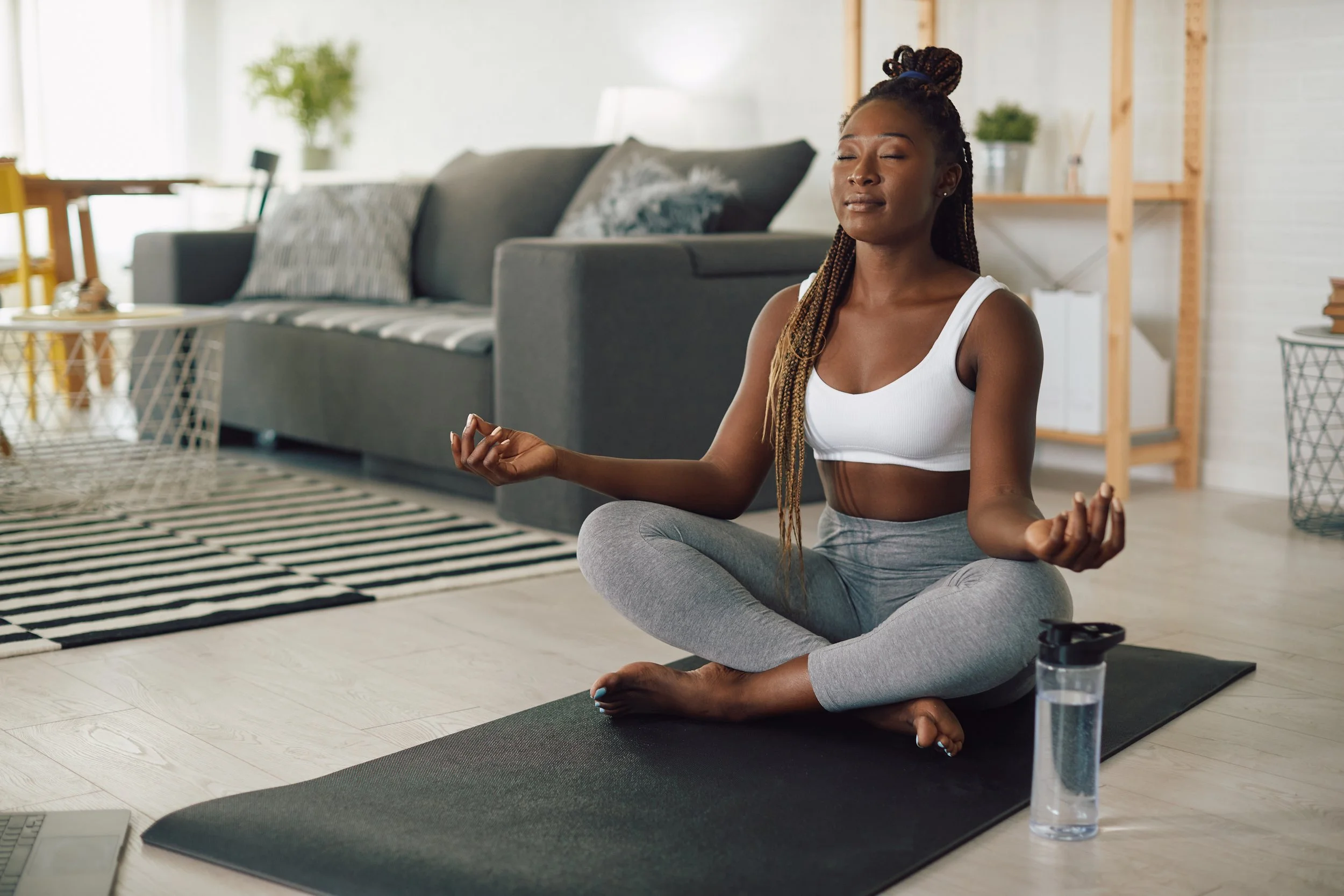 Woman meditating in a living room, sitting cross-legged on a yoga mat, with eyes closed and hands in a mudra position. A water bottle is placed next to her on the floor.