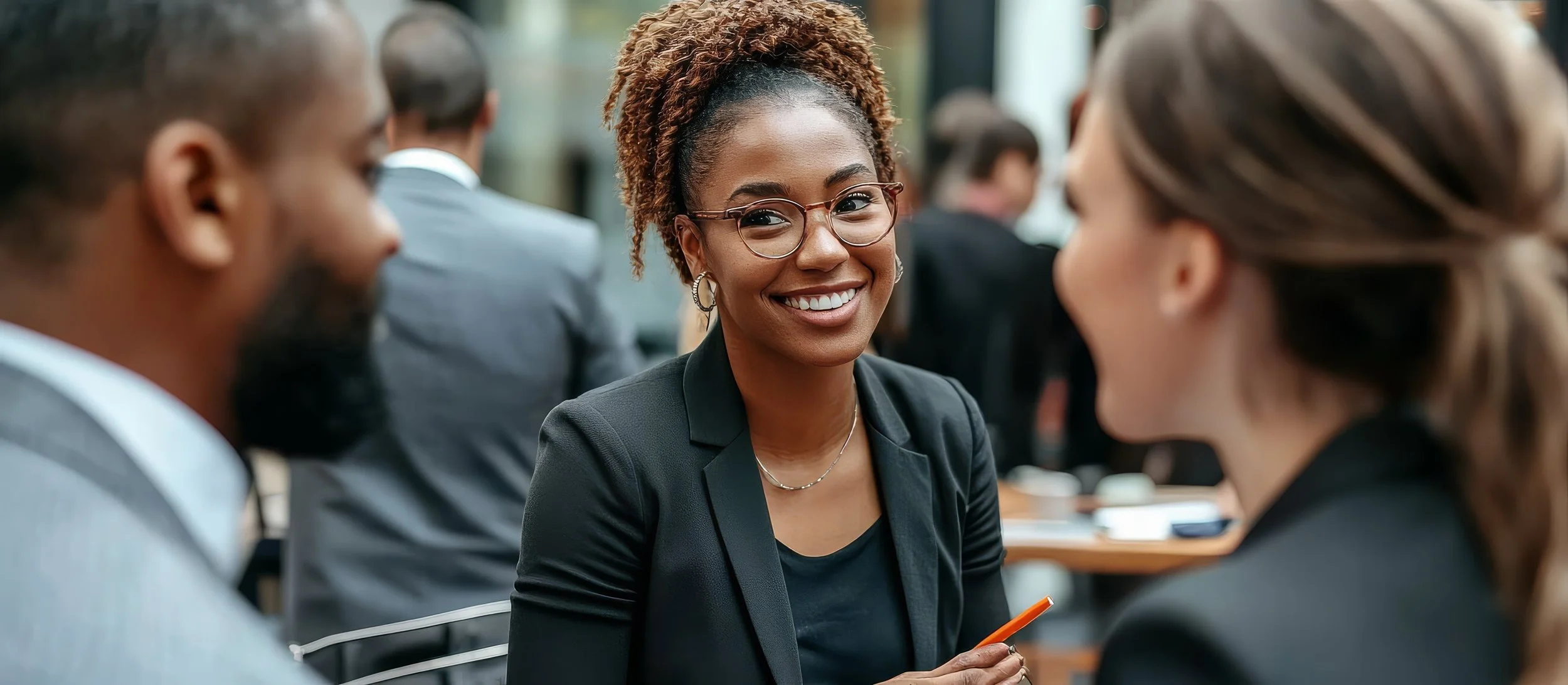 A woman with glasses smiling and engaging in conversation, surrounded by people in business attire at a professional event.