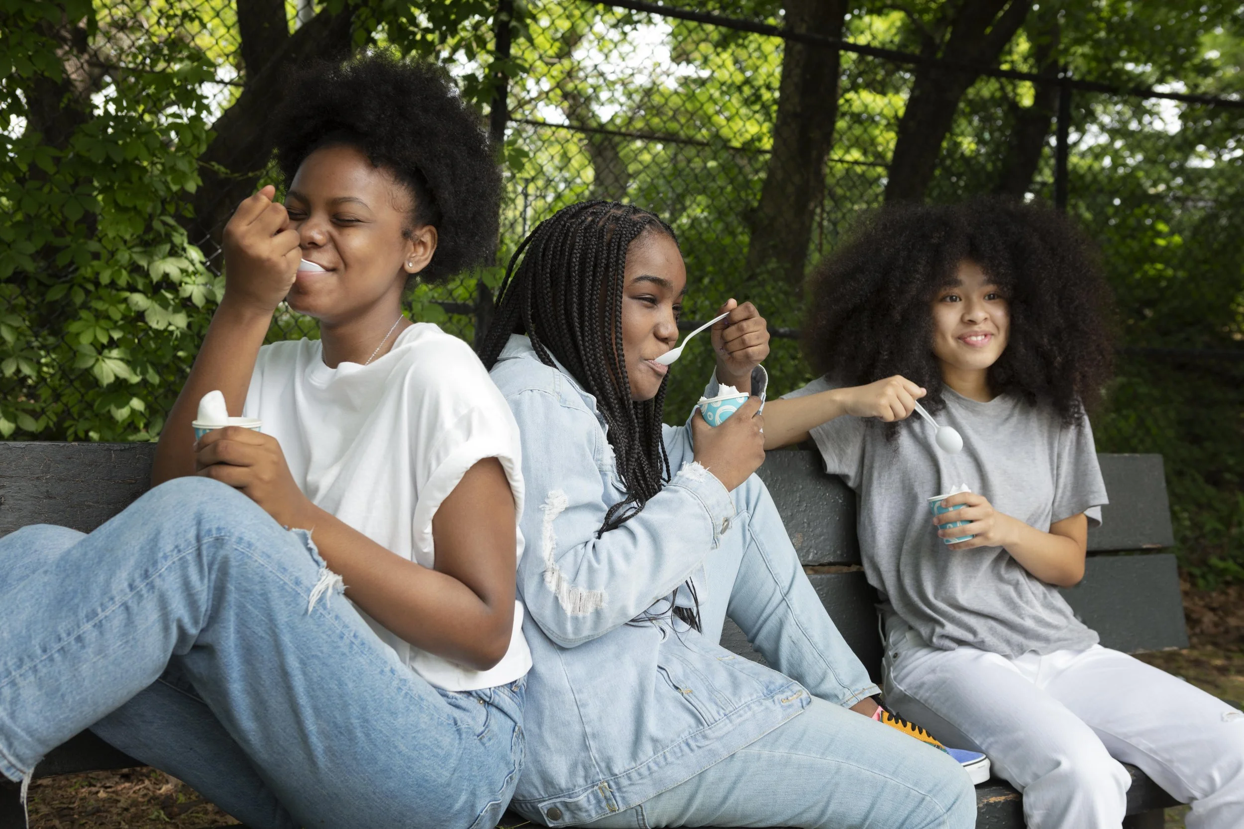 Three young women sitting on a park bench eating ice cream and smiling, with trees and a fence in the background.