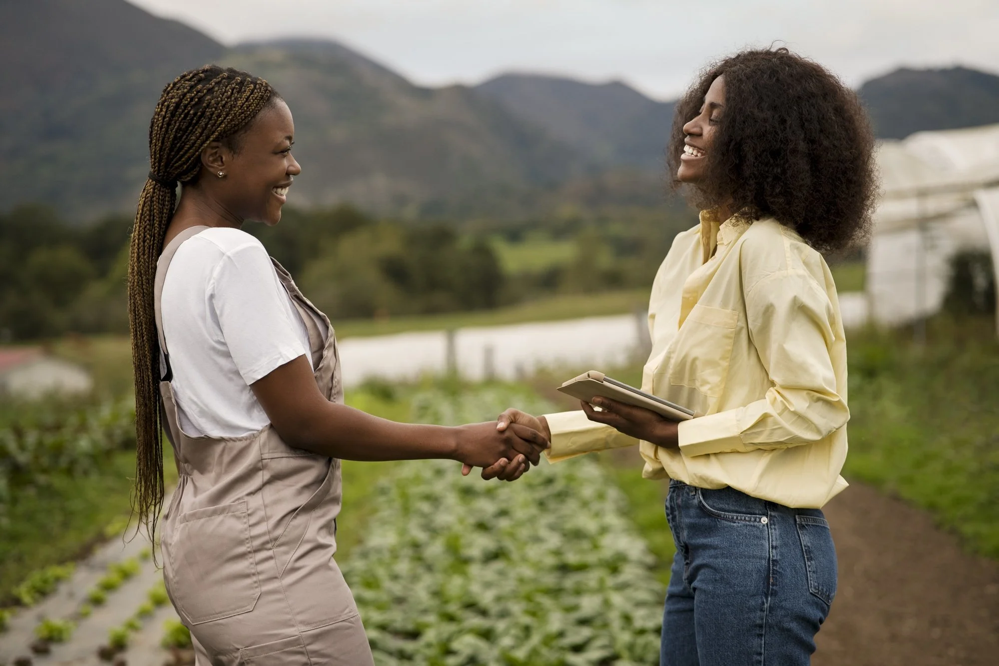 Two women smiling and shaking hands in a farm field, one holding a digital tablet.