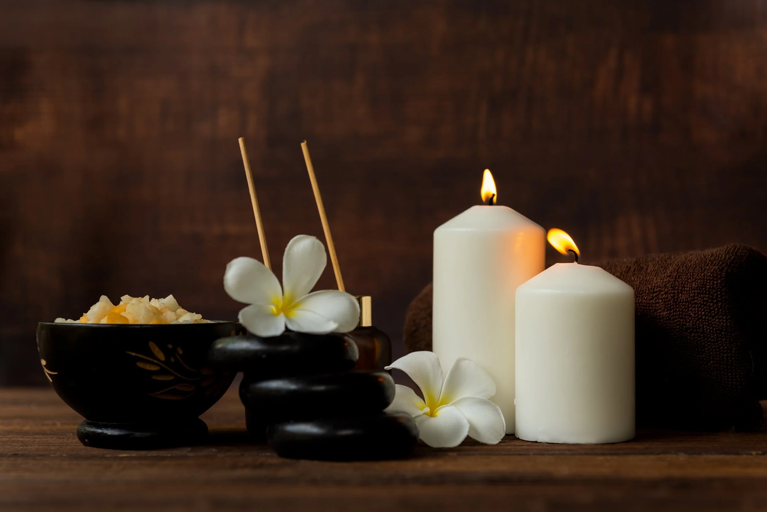 Spa setting with lit candles, white flowers, black stones, bowl of bath salts, and incense sticks on a wooden surface.