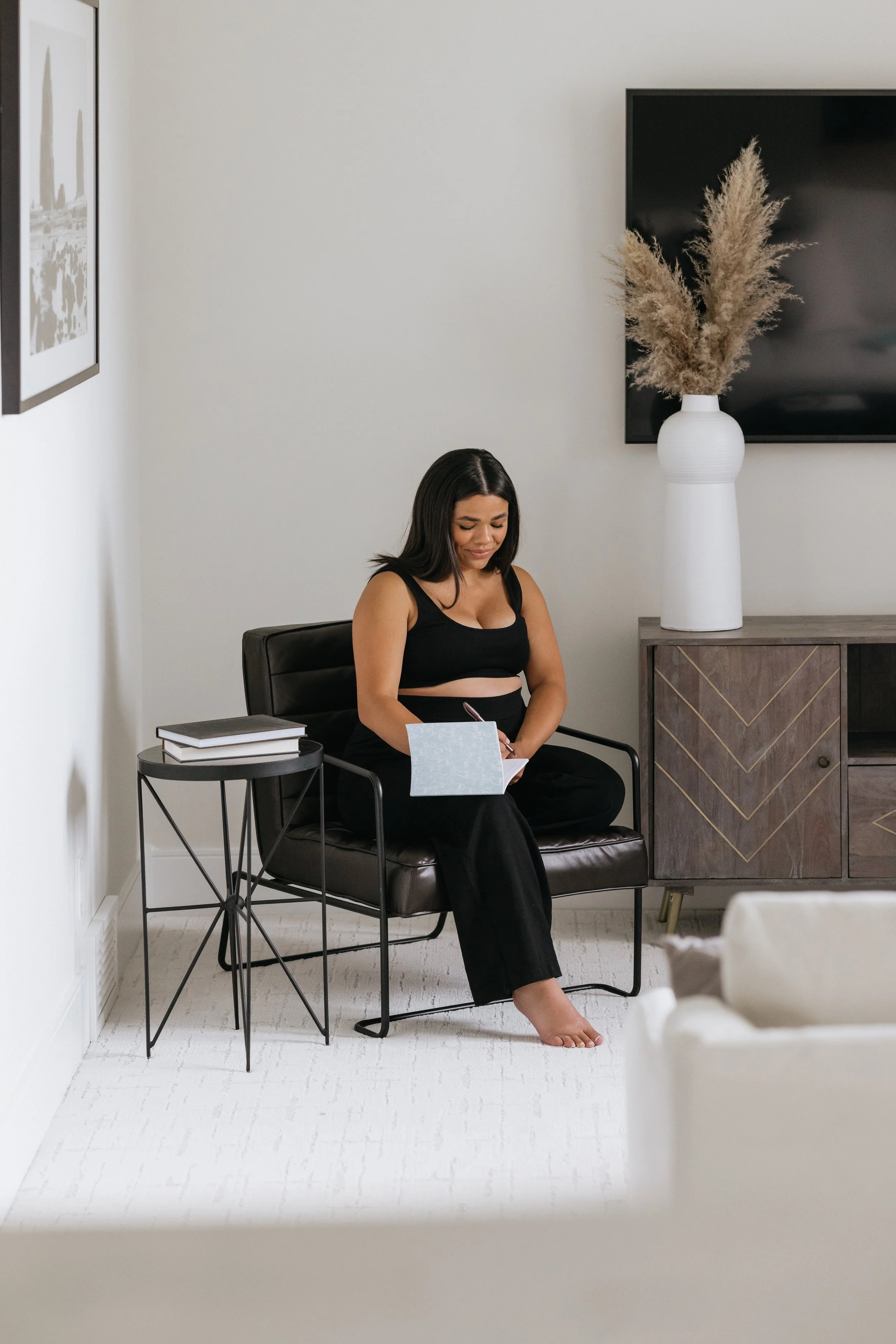 Woman sitting in a modern living room, writing in a notebook. She is seated on a black chair next to a side table with books and a decorative vase on a cabinet nearby.