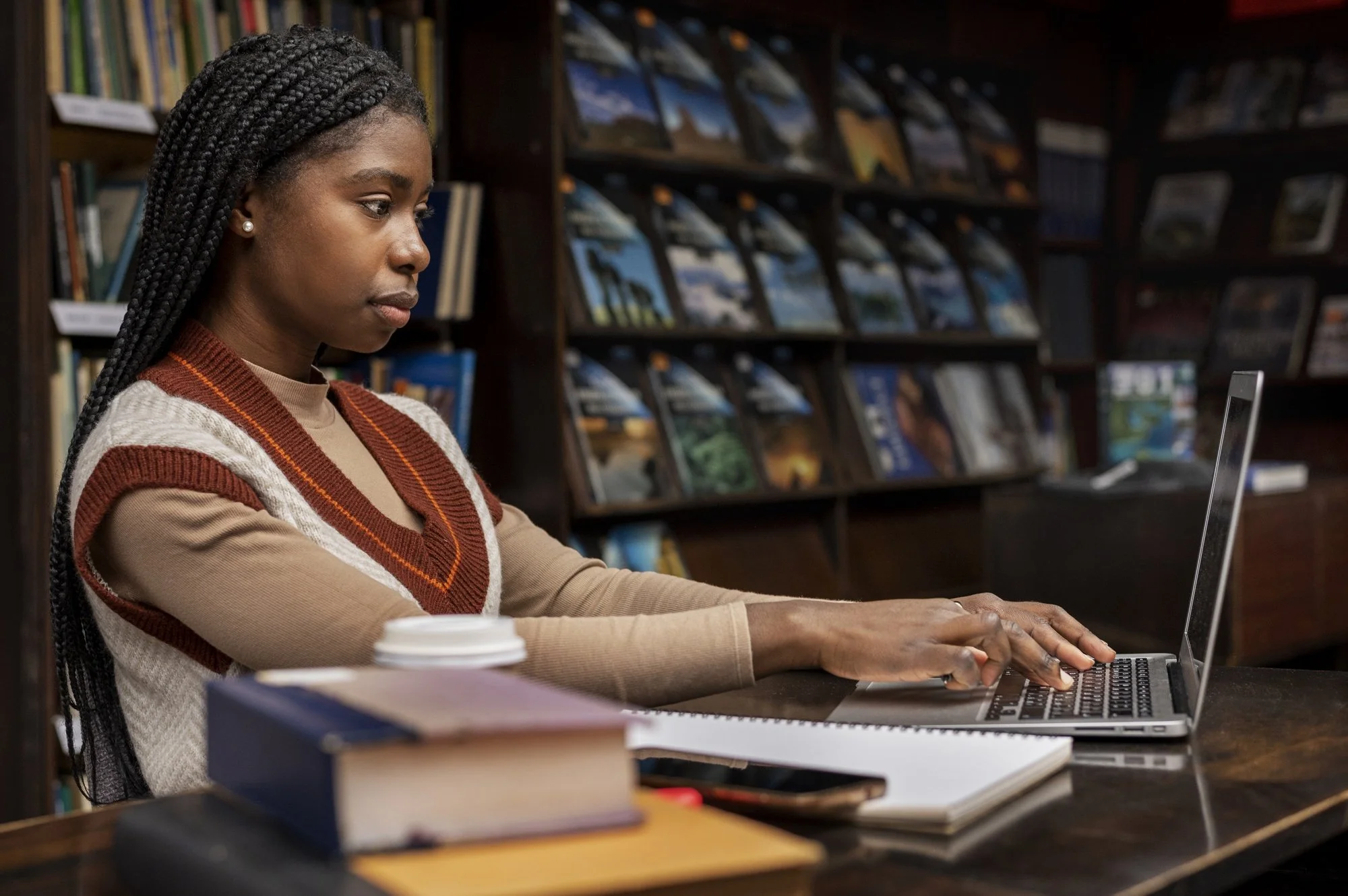 Woman with braids working on a laptop in a library, surrounded by books and notebooks.