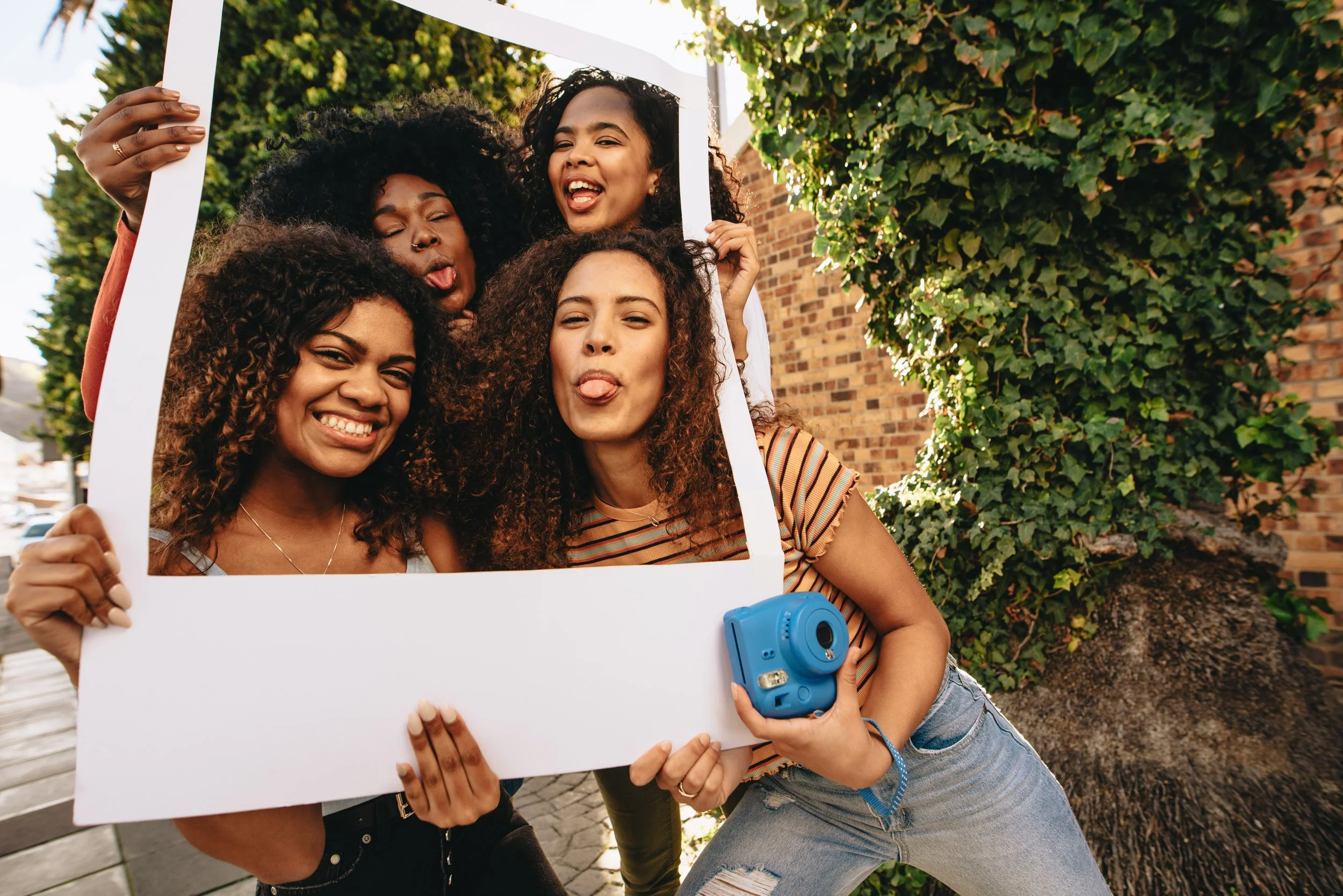 Four young women smiling and posing for a photo with a large white frame, one holding a blue instant camera, outdoors.