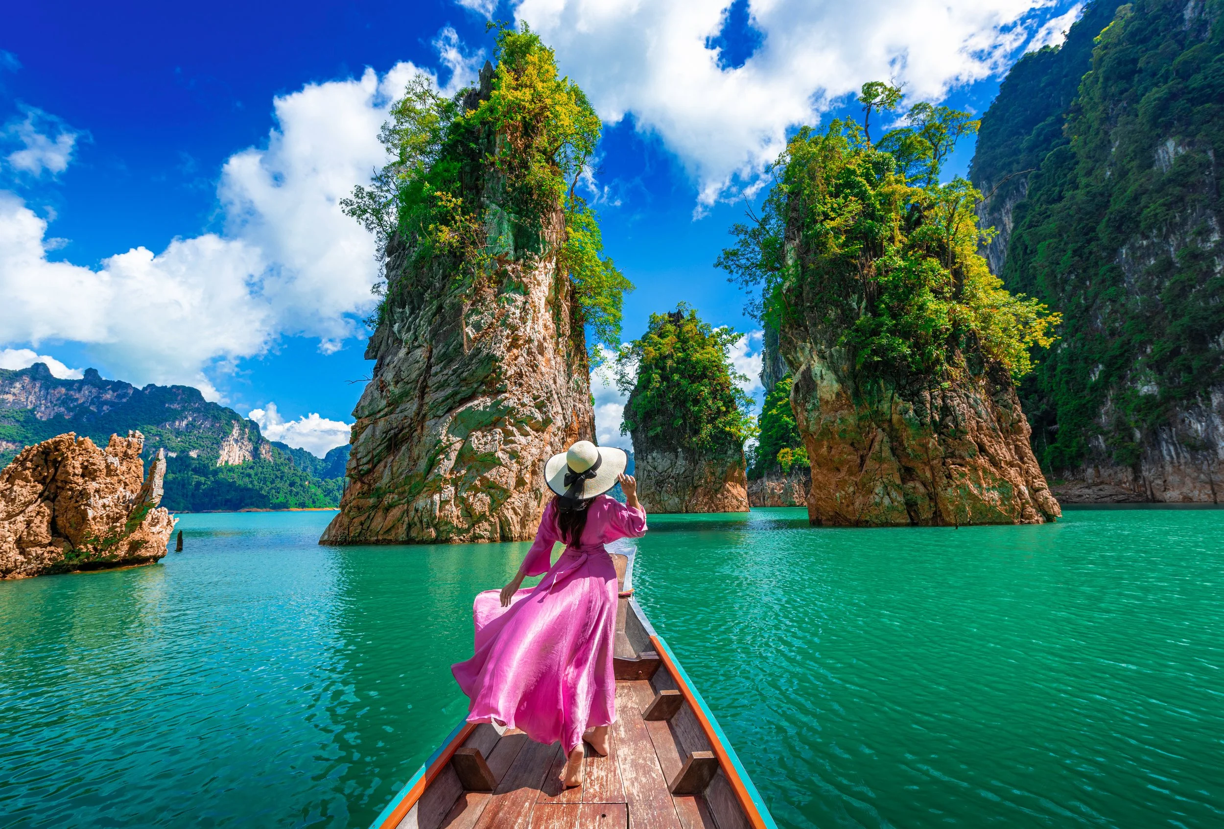 Woman in pink dress on boat at Cheow Lan Lake, surrounded by limestone karsts and lush greenery, under a bright blue sky.