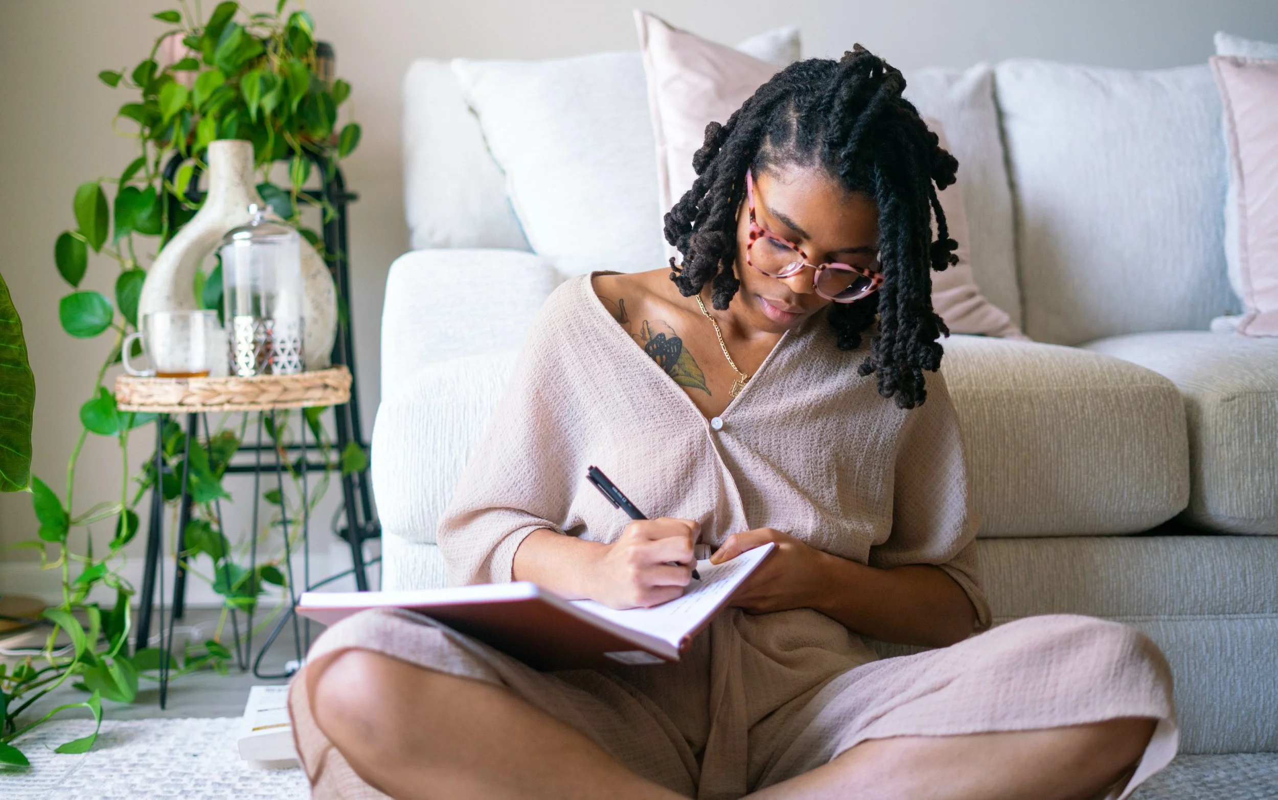 Person sitting on the floor writing in a notebook, surrounded by plants and a cozy sofa.