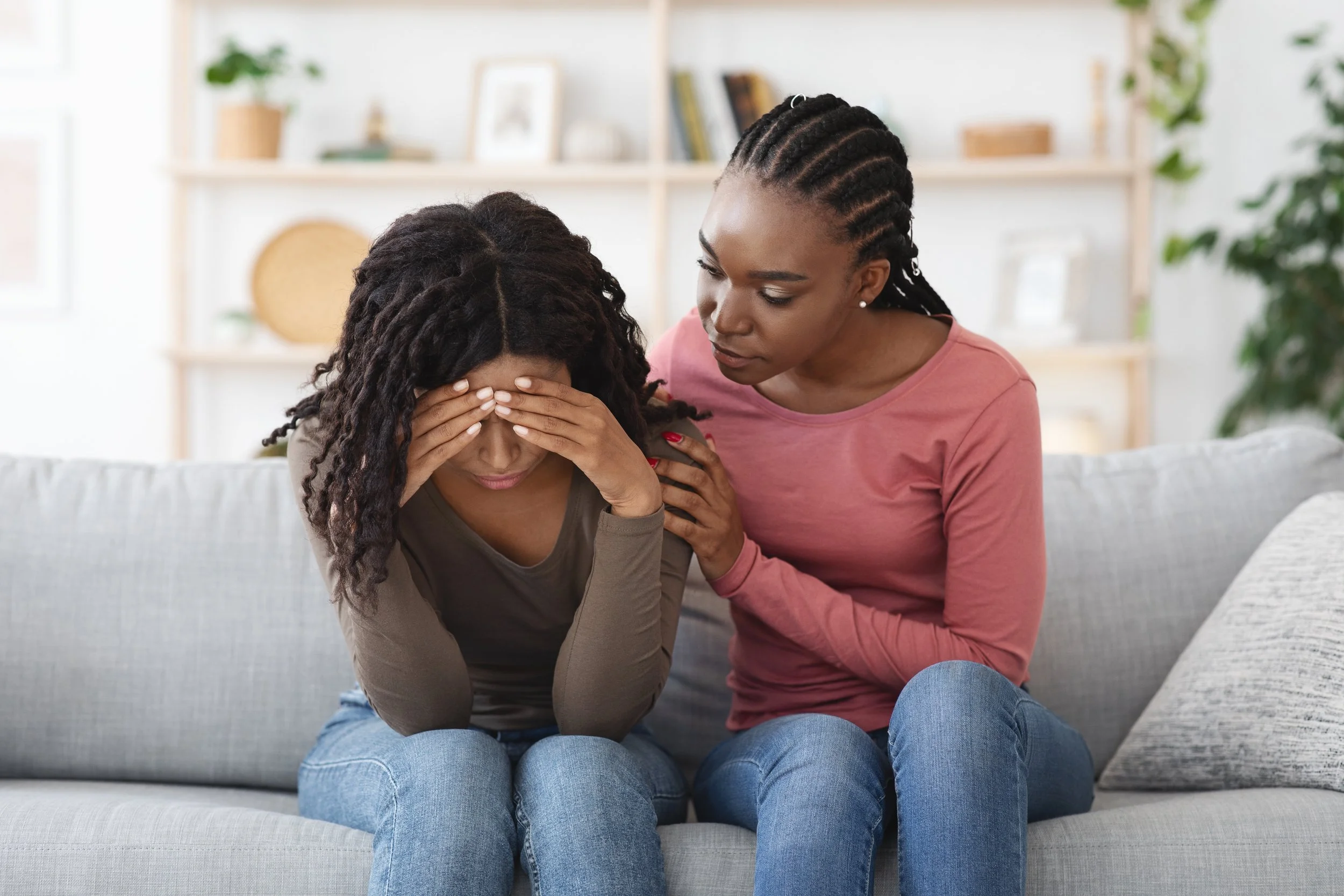 Two women sitting on a couch; one appears upset with her face in her hands, and the other is comforting her with a hand on her shoulder.
