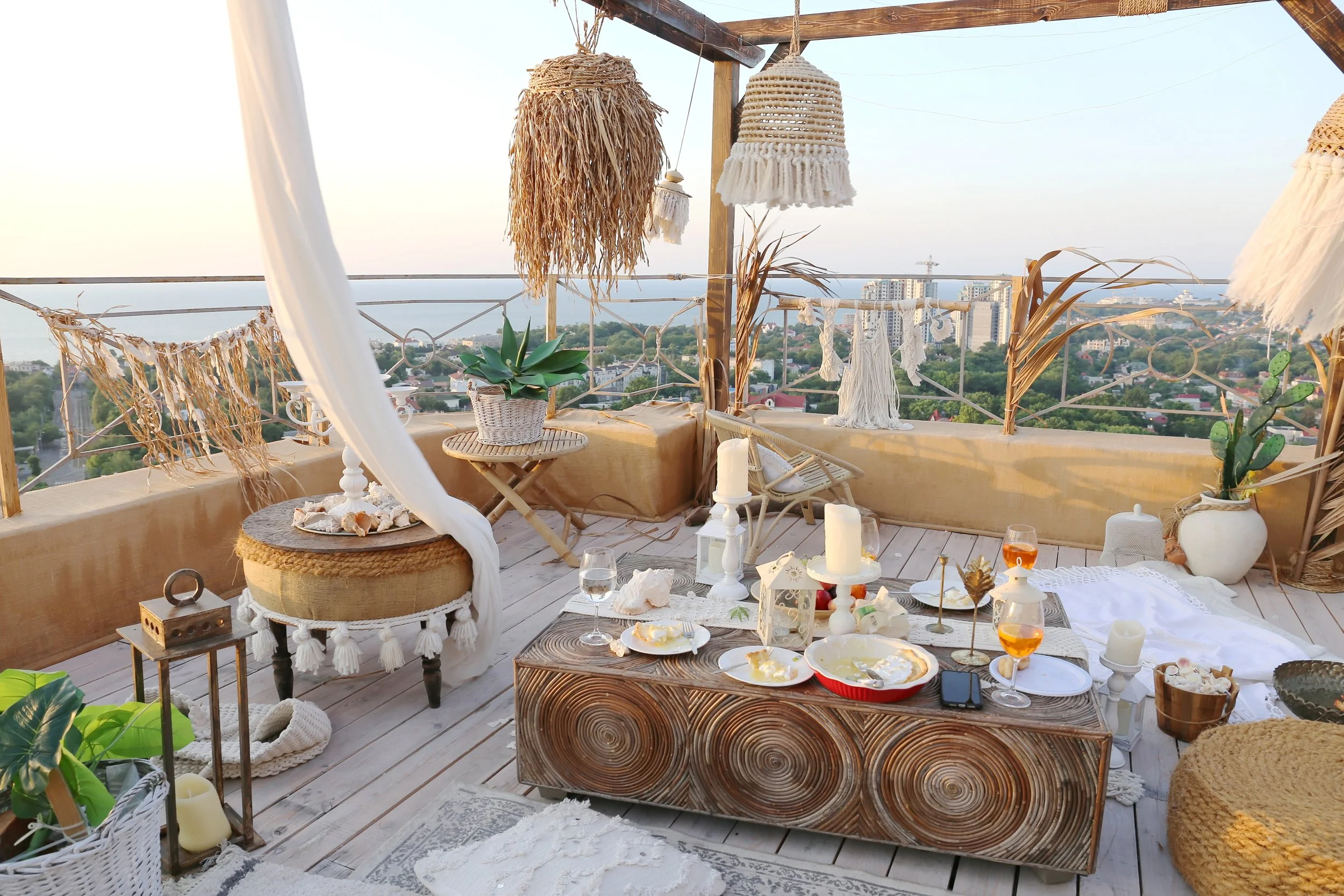 Bohemian style rooftop terrace with wooden furniture, decorative lanterns, candles, and potted plants overlooking a coastal cityscape.