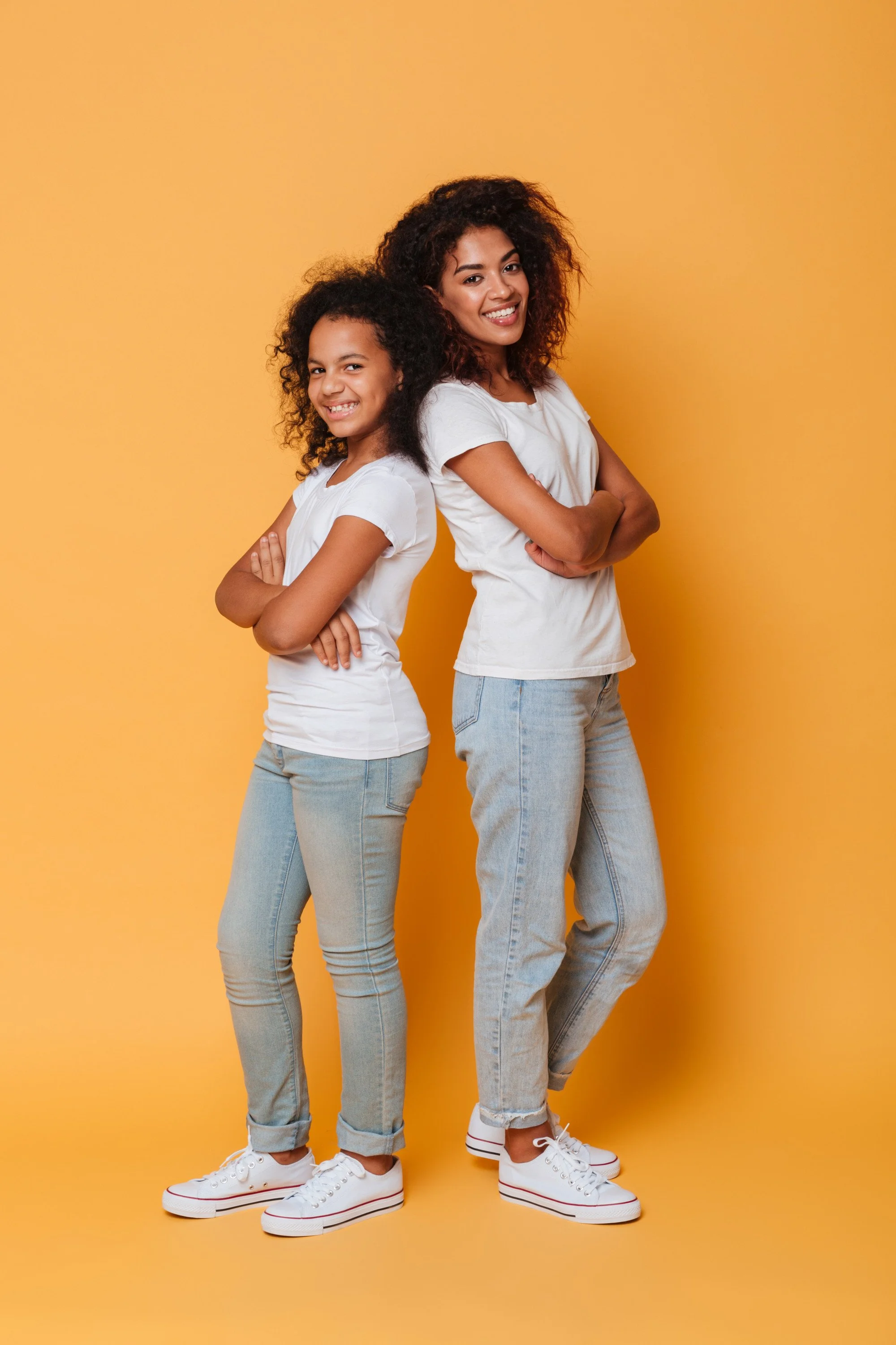 Two people smiling, standing back to back, wearing white t-shirts and jeans on a yellow background.
