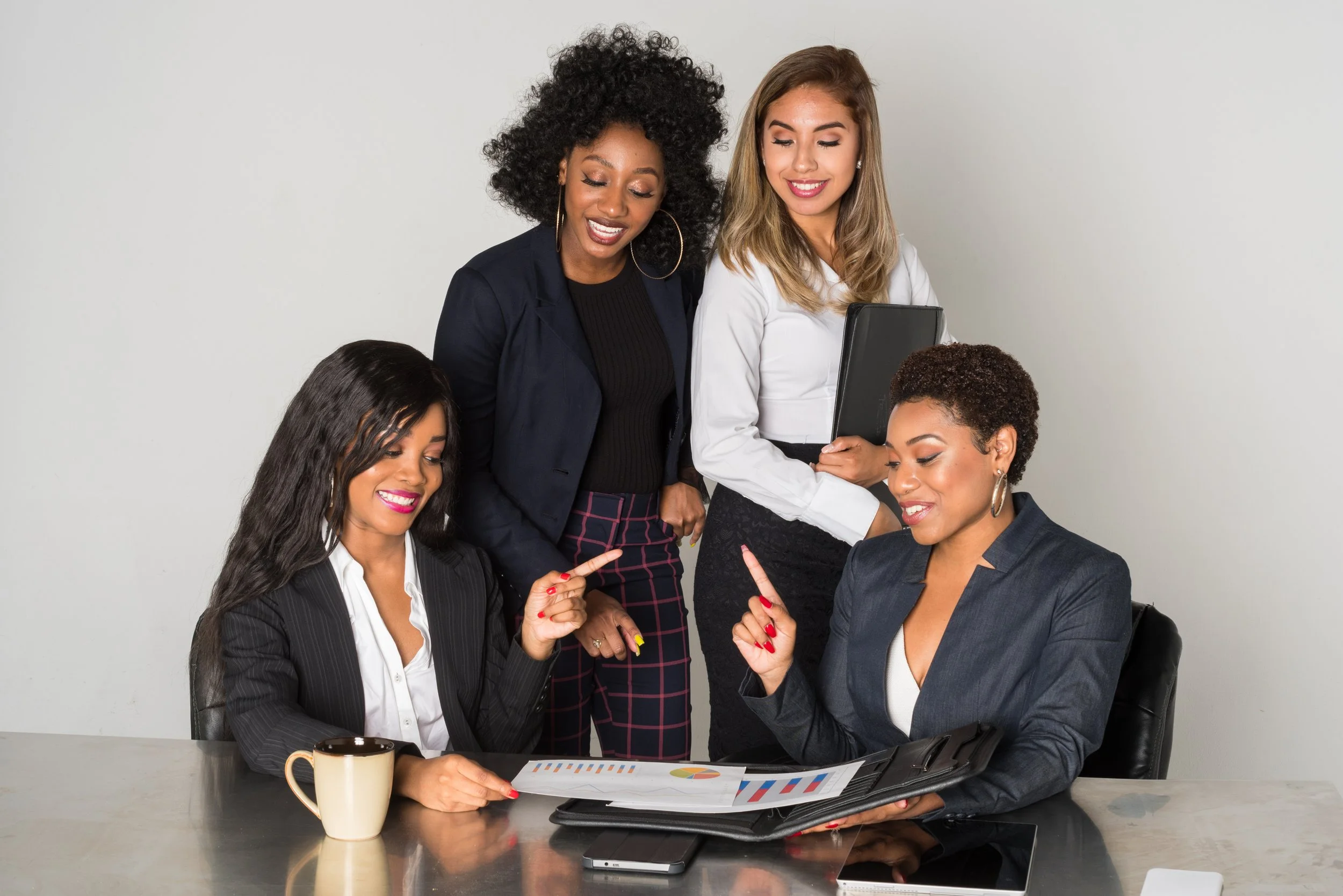 Four women in business attire engaged in a discussion around a table with documents, a coffee mug, and electronic devices.