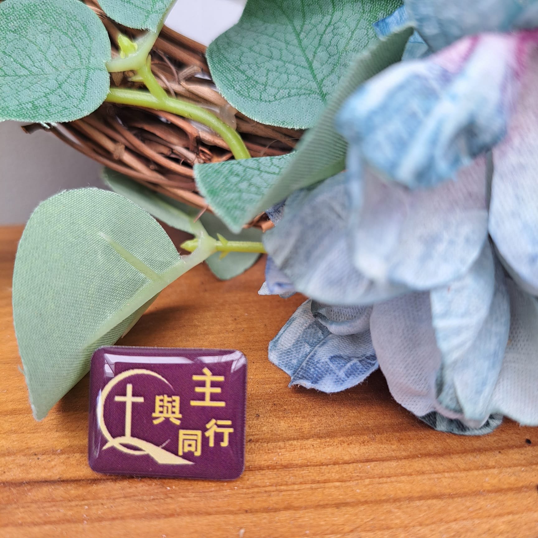A close-up of a wooden surface with an artificial flower arrangement and a Chinese-character pin featuring a cross, symbols of faith.