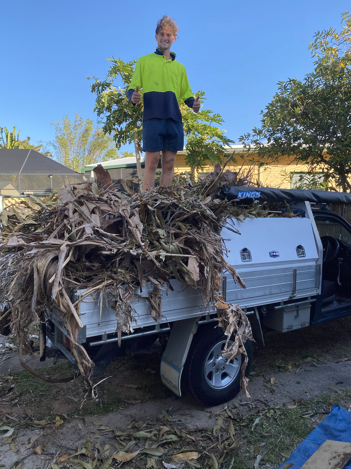 A young man in a bright yellow-green jacket and dark shorts standing on a small truck bed filled with dry, dead leaves and plant debris, giving a thumbs-up. The background shows a clear blue sky, trees, and a house.
