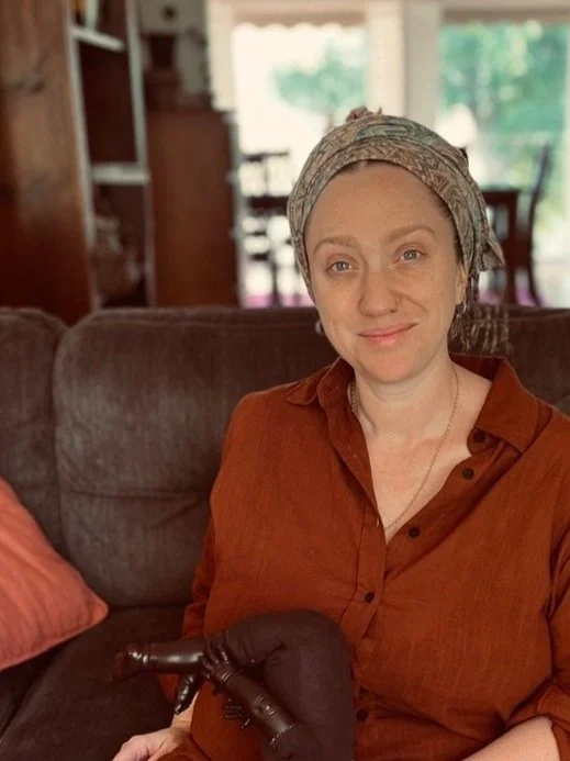 A midwife sitting on a couch with a brown shirt and headscarf, in a living room setting.