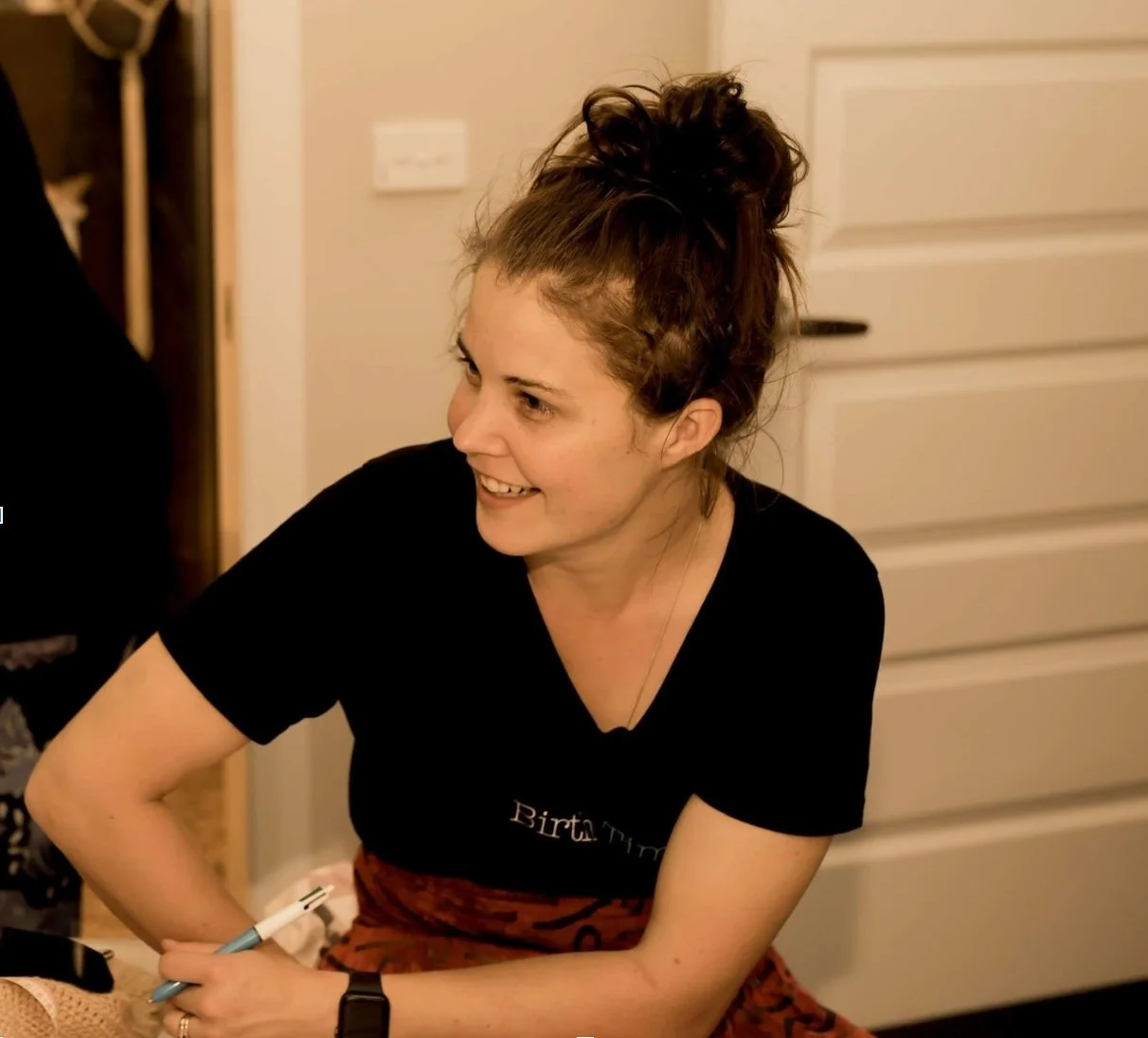 A midwife smiling and sitting near a door, holding a pen.