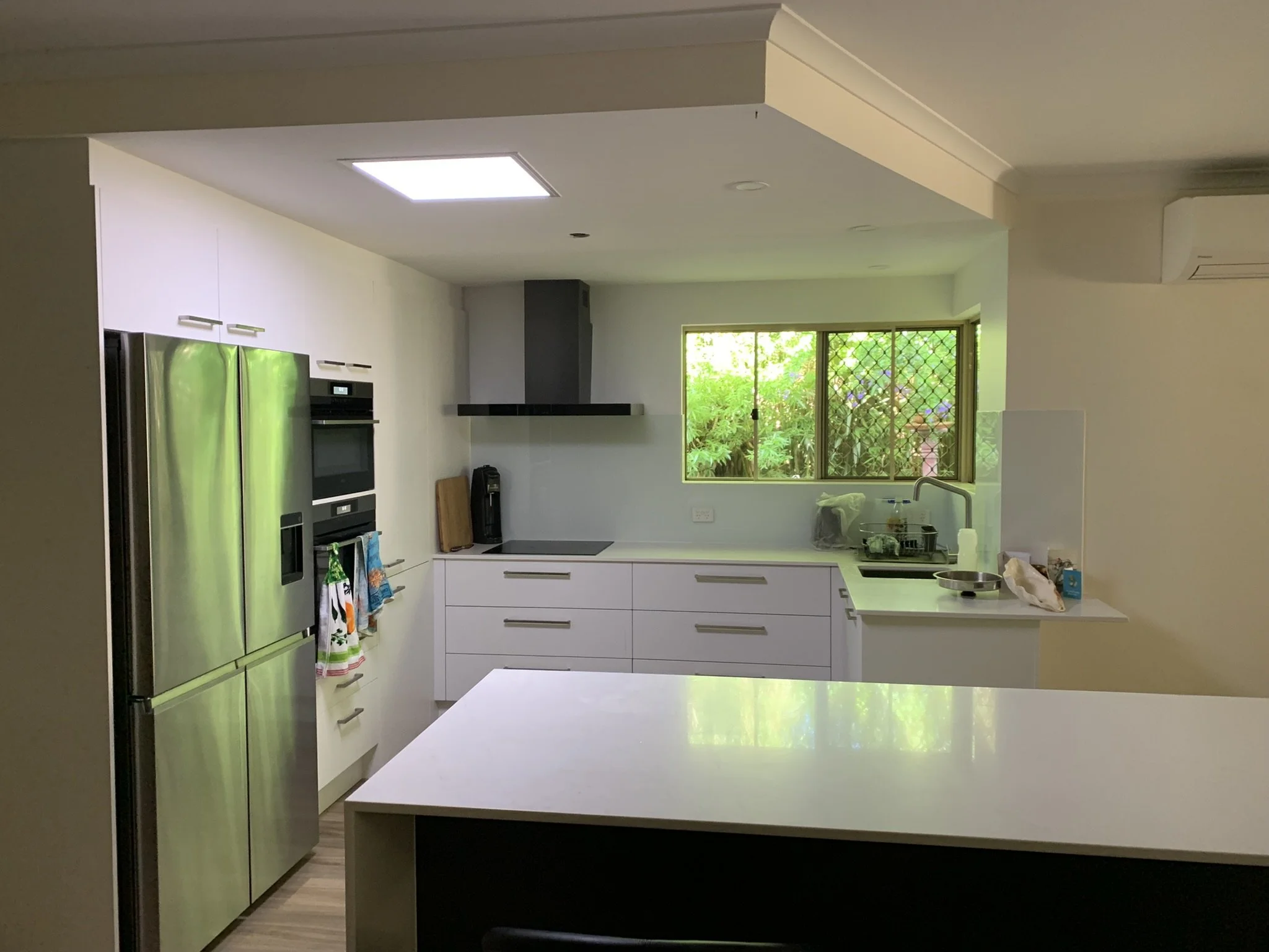 Modern kitchen with an illume skylight, stainless steel refrigerator, built-in oven, microwave, white cabinets, island counter, and window view.