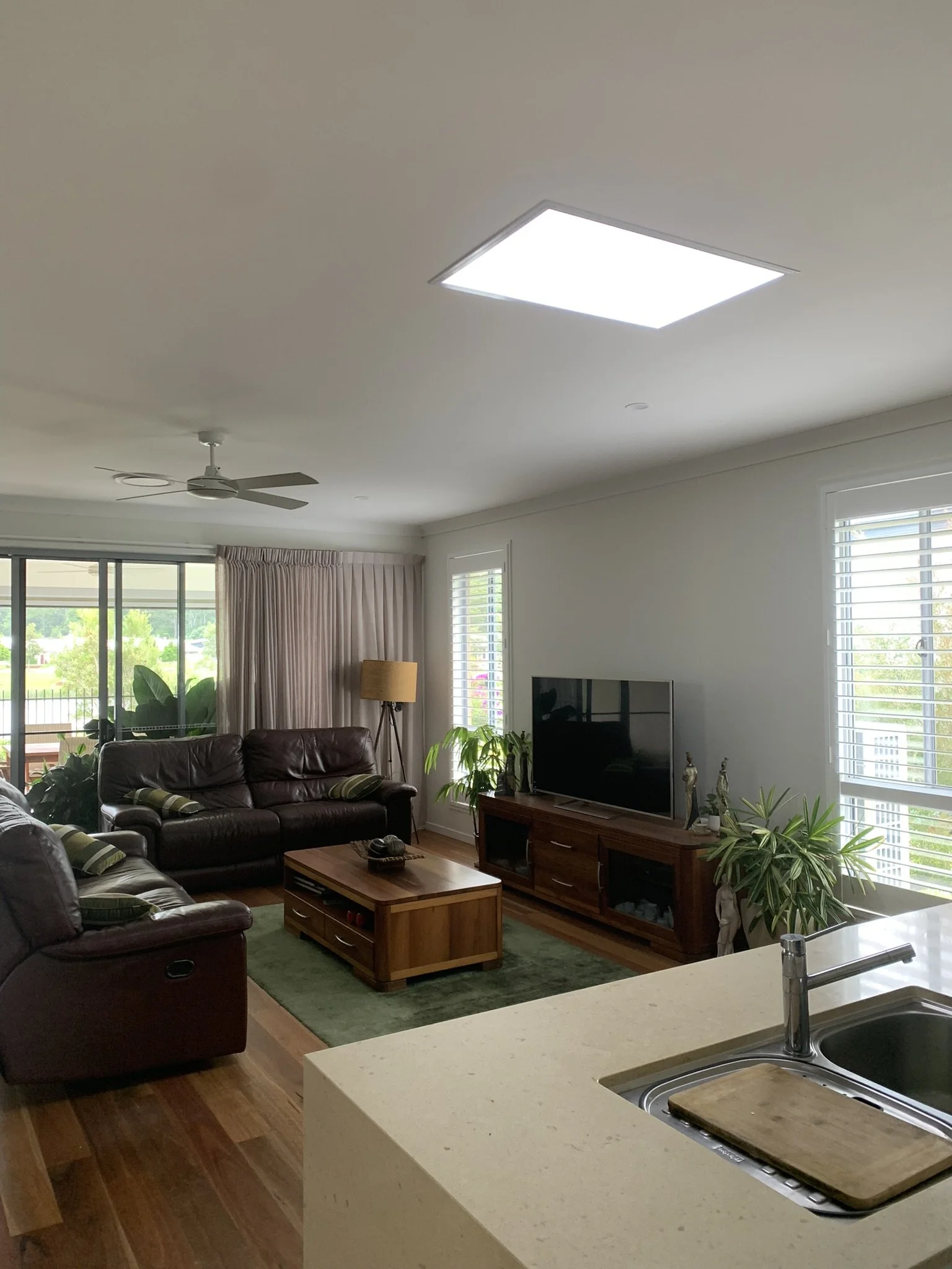Modern living room with leather couches, a wooden coffee table, TV on a cabinet, large windows with shutters, and green plants. Kitchen sink and countertop in foreground. The square illume skylight is giving a modern look te o the room.