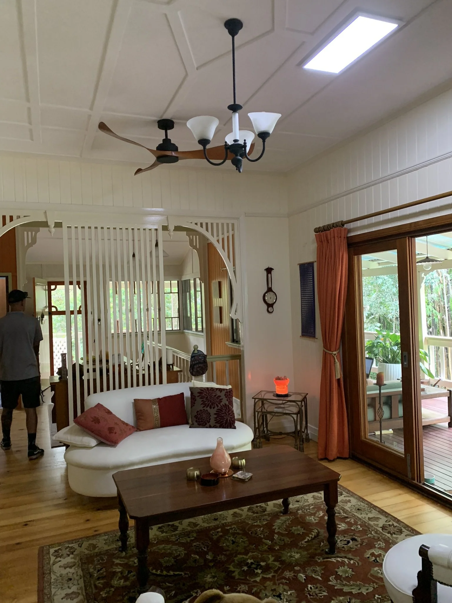 Living room interior with a white sofa, decorative pillows, wooden coffee table, ceiling fan, and chandelier. Sliding glass doors lead to a deck. The room features an illume skylight, wooden floors, and decorative elements like wall art.